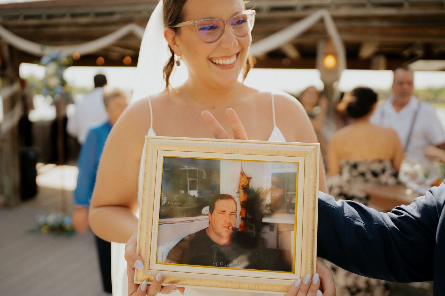 Smiling bride in veil holding framed photo of man at outdoor wedding reception.