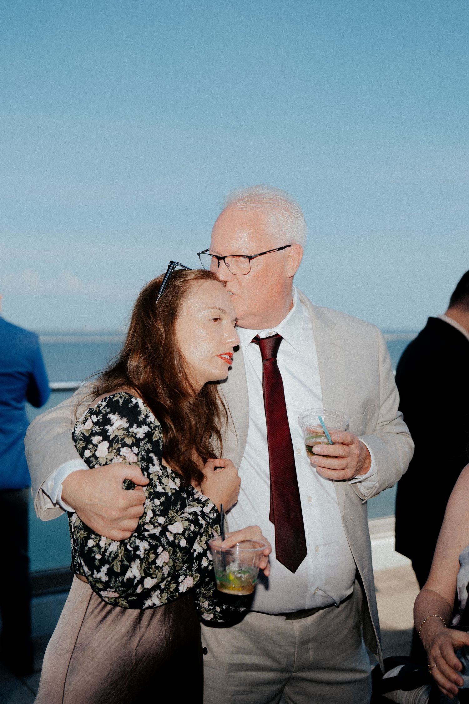 Older gentleman in beige suit and burgundy tie embracing a young woman at an outdoor wedding reception.