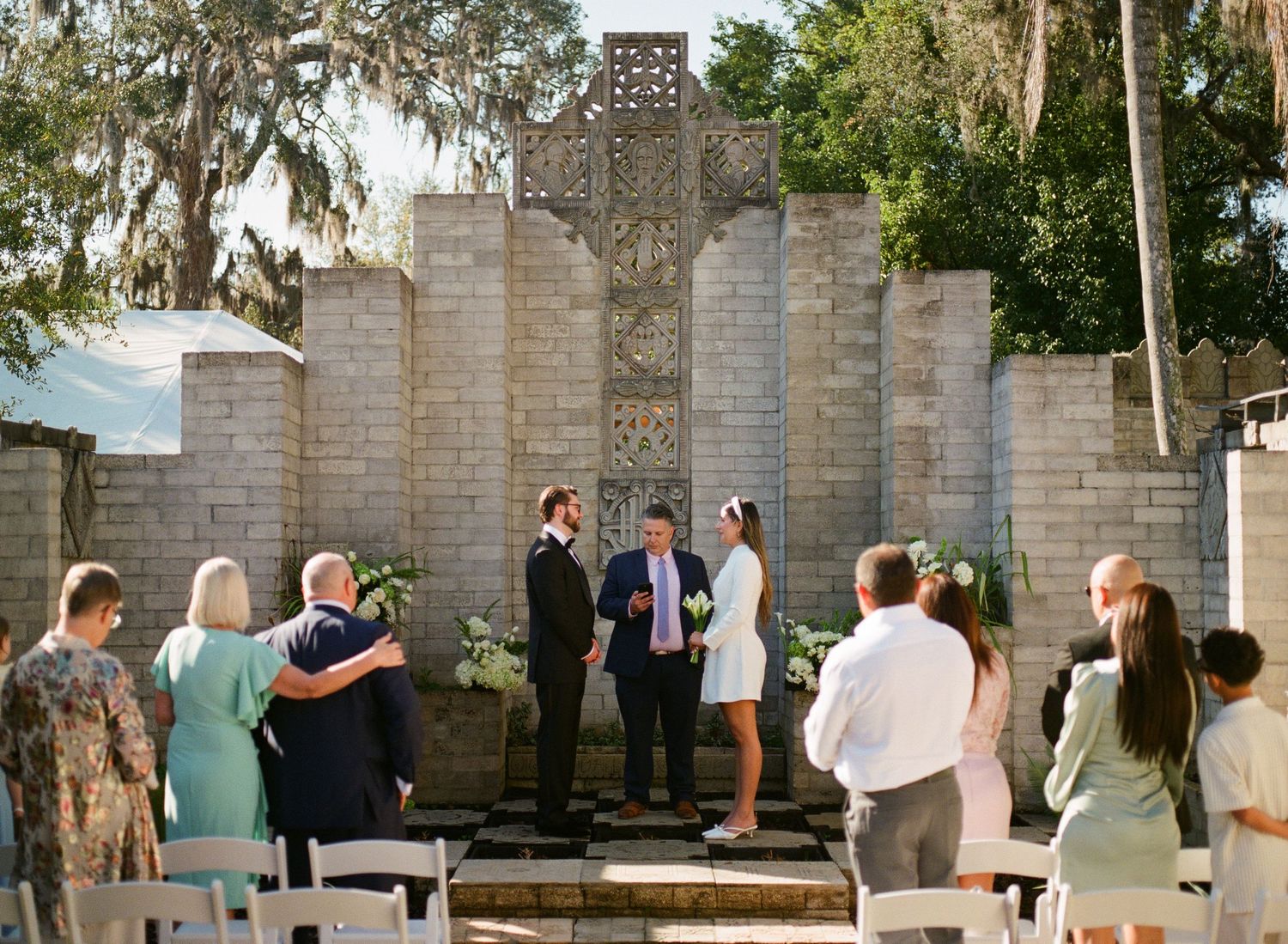 Intimate outdoor wedding ceremony with guests gathered before an ornate stone cross altar backdrop.