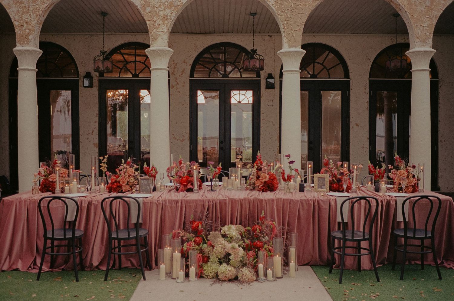 Romantic outdoor wedding reception with long pink-draped tables, red floral arrangements under arched colonnade.
