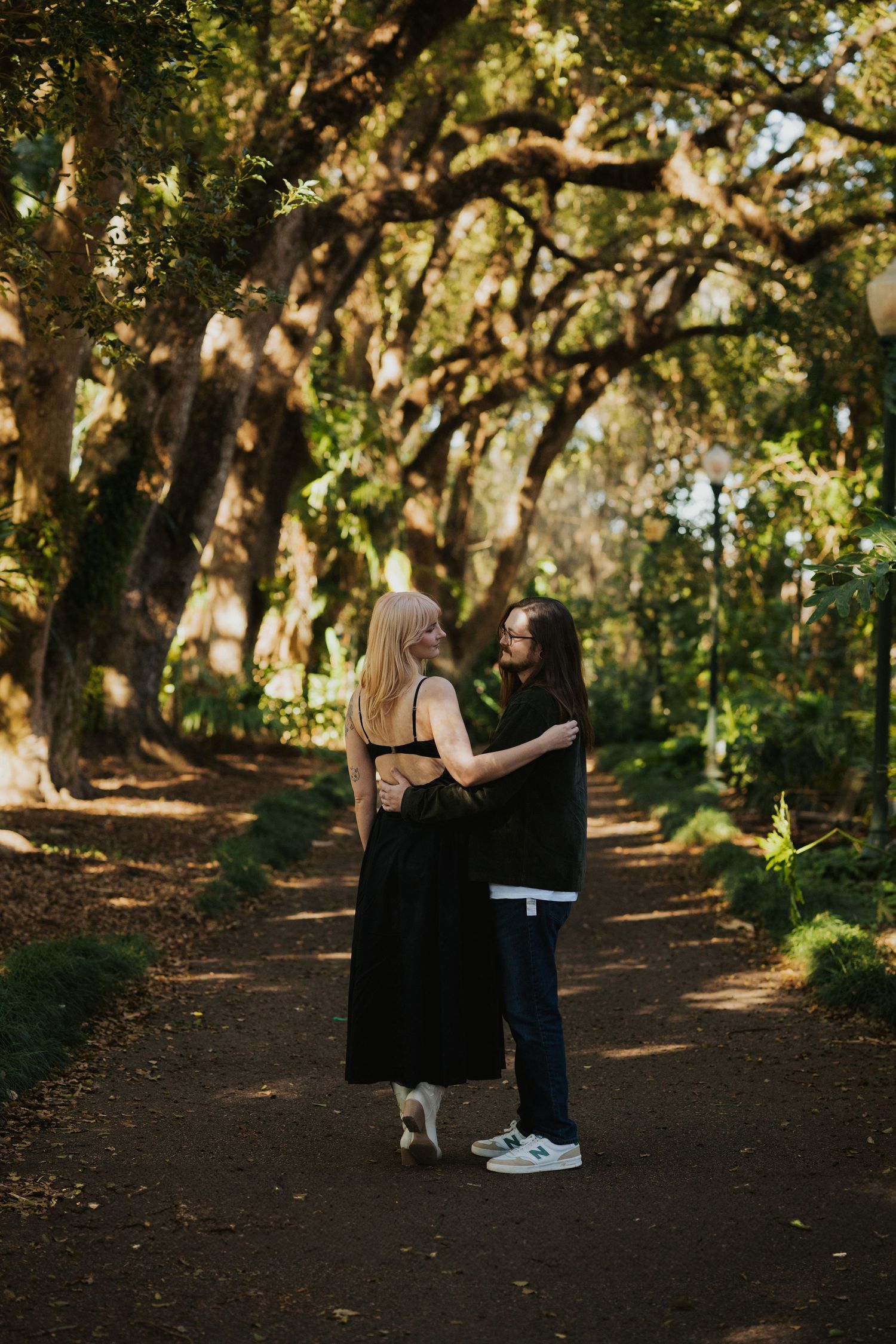 A couple embraces on a tree-lined dirt path in a sunlit forest.