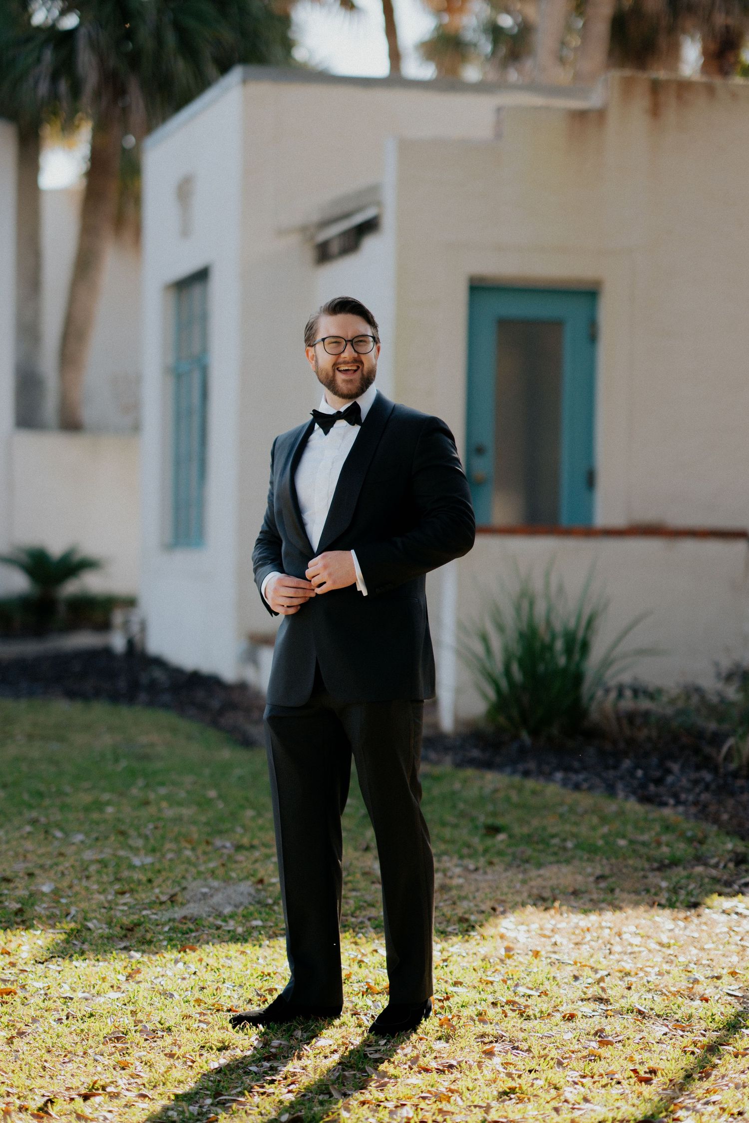 A groom in a classic black tuxedo and bow tie poses outdoors in front of a white building with palm trees.