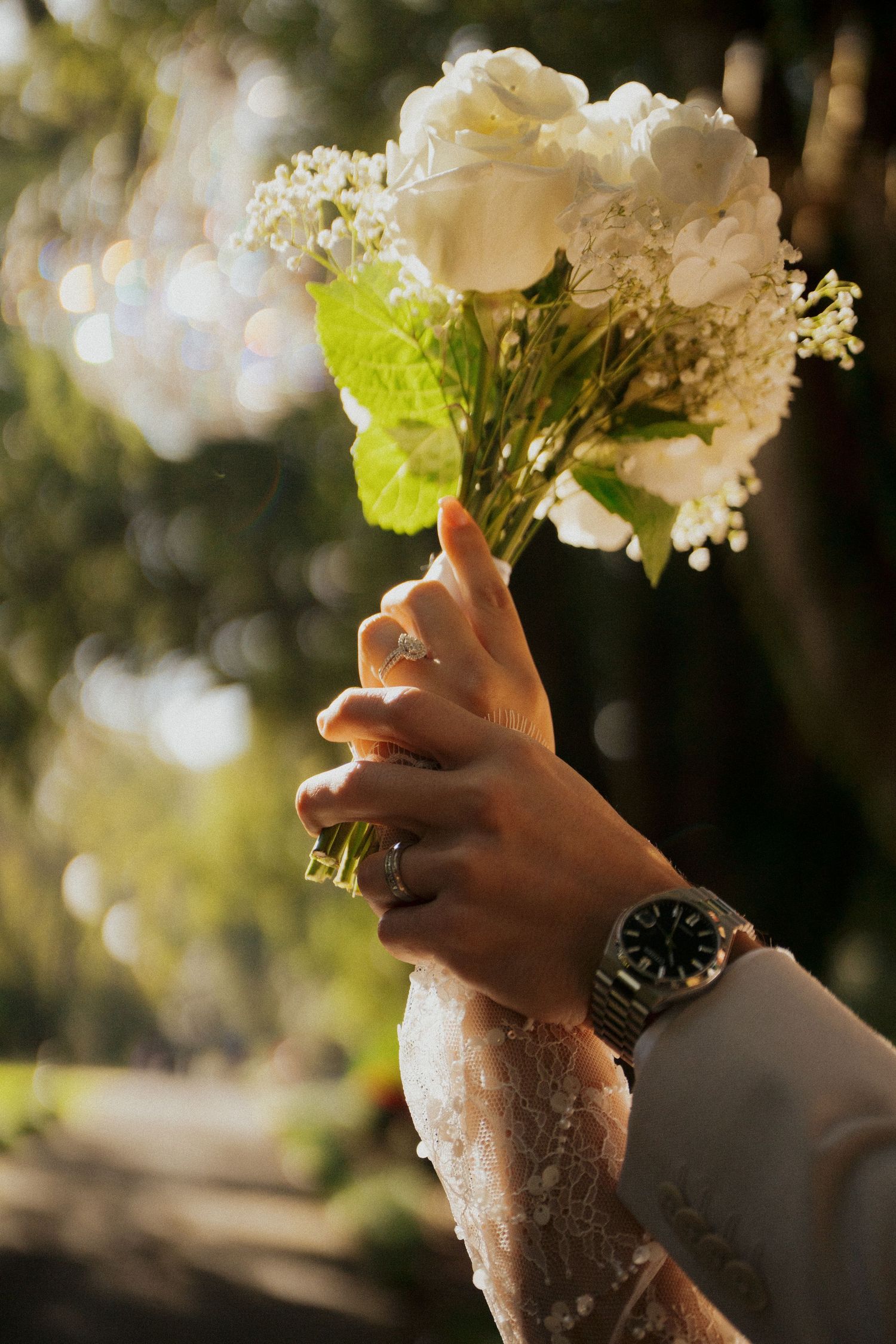 Hands holding a white and green floral bouquet against a blurred outdoor background with sunlight filtering through.