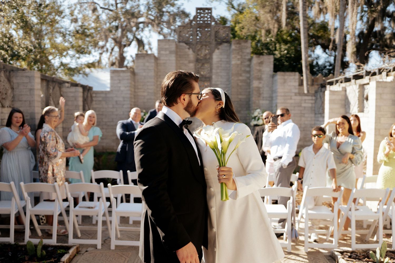 Romantic wedding ceremony in front of a stone wall backdrop with guests seated on white chairs.
