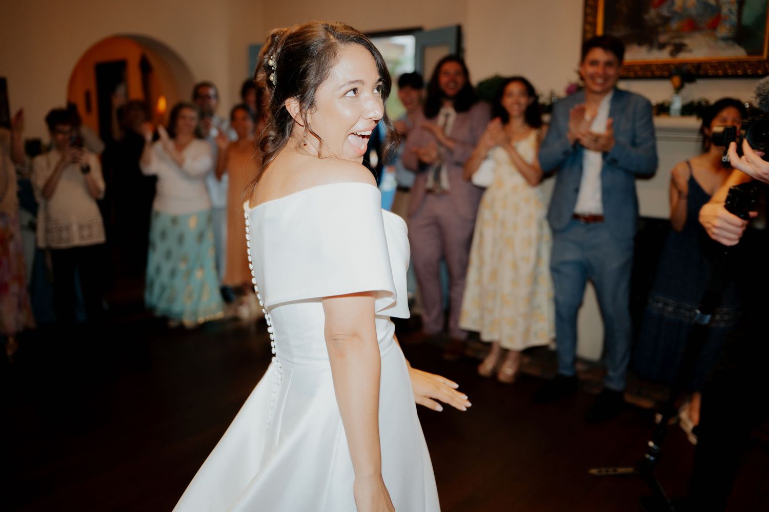 A bride in an elegant off-shoulder white wedding dress turns to look back at wedding guests during a reception.