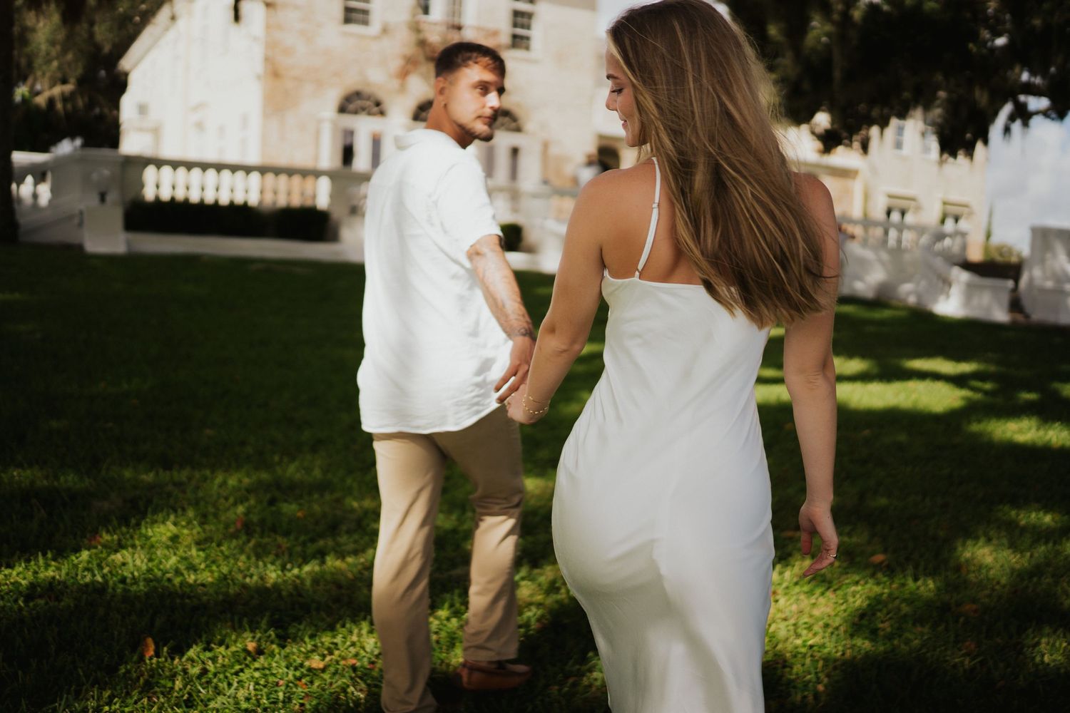 Couple in light summer wedding attire walks together through a lush green garden setting.