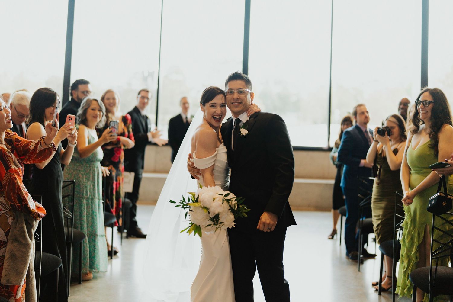 Wedding guests gather in a bright modern venue with floor-to-ceiling windows and white curtains for a ceremony.