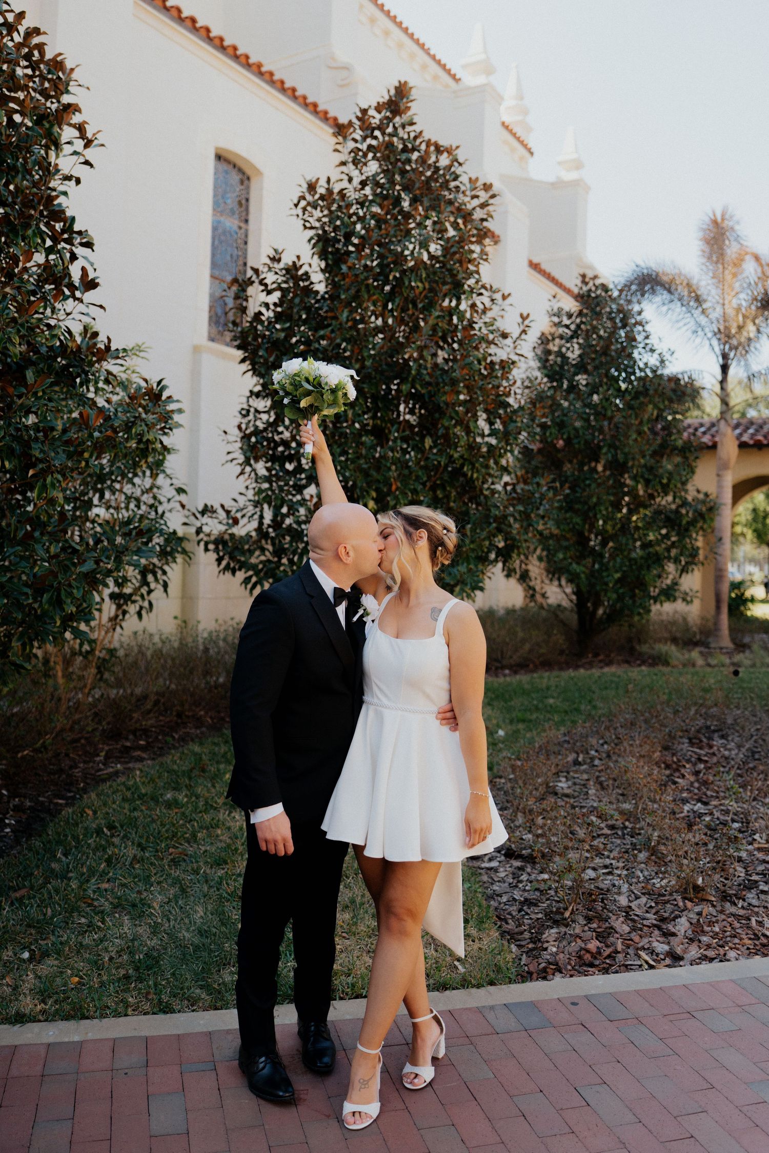 Bride and groom share a kiss outdoors while bride raises white bouquet in the air.