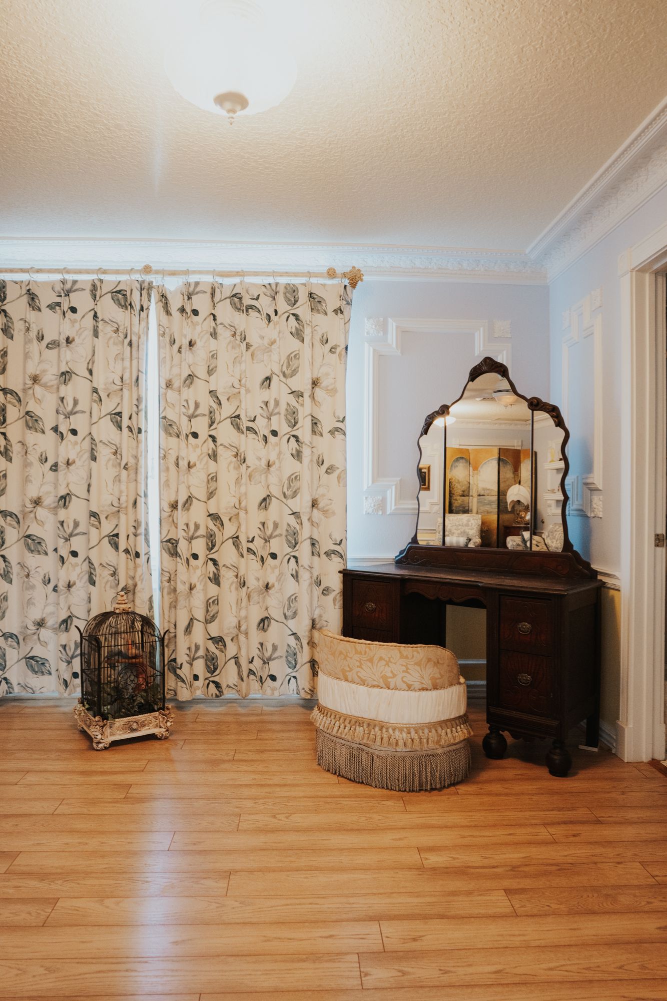 Vintage inspired room with floral curtains, antique vanity, and decorative seating on hardwood floors.