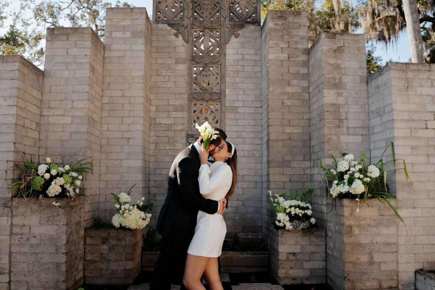 A romantic couple shares a tender moment against a decorative stone wall adorned with white floral arrangements.