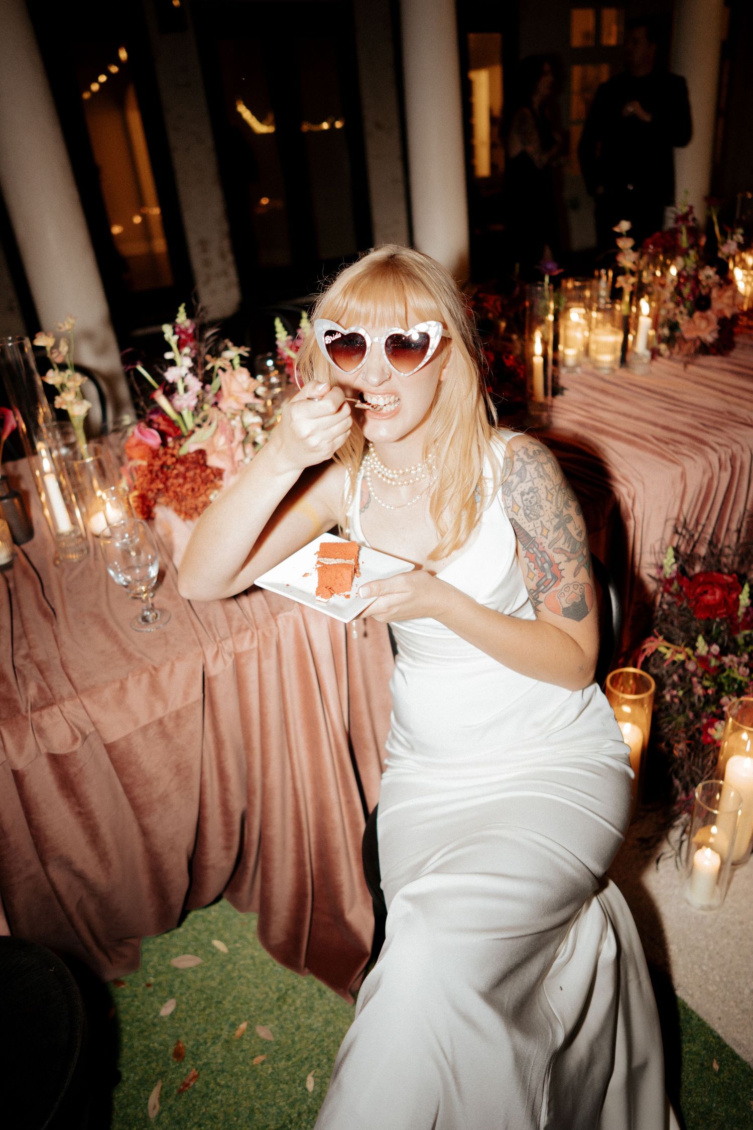 Tattooed bride in white gown wearing heart sunglasses enjoys cake at candlelit reception.