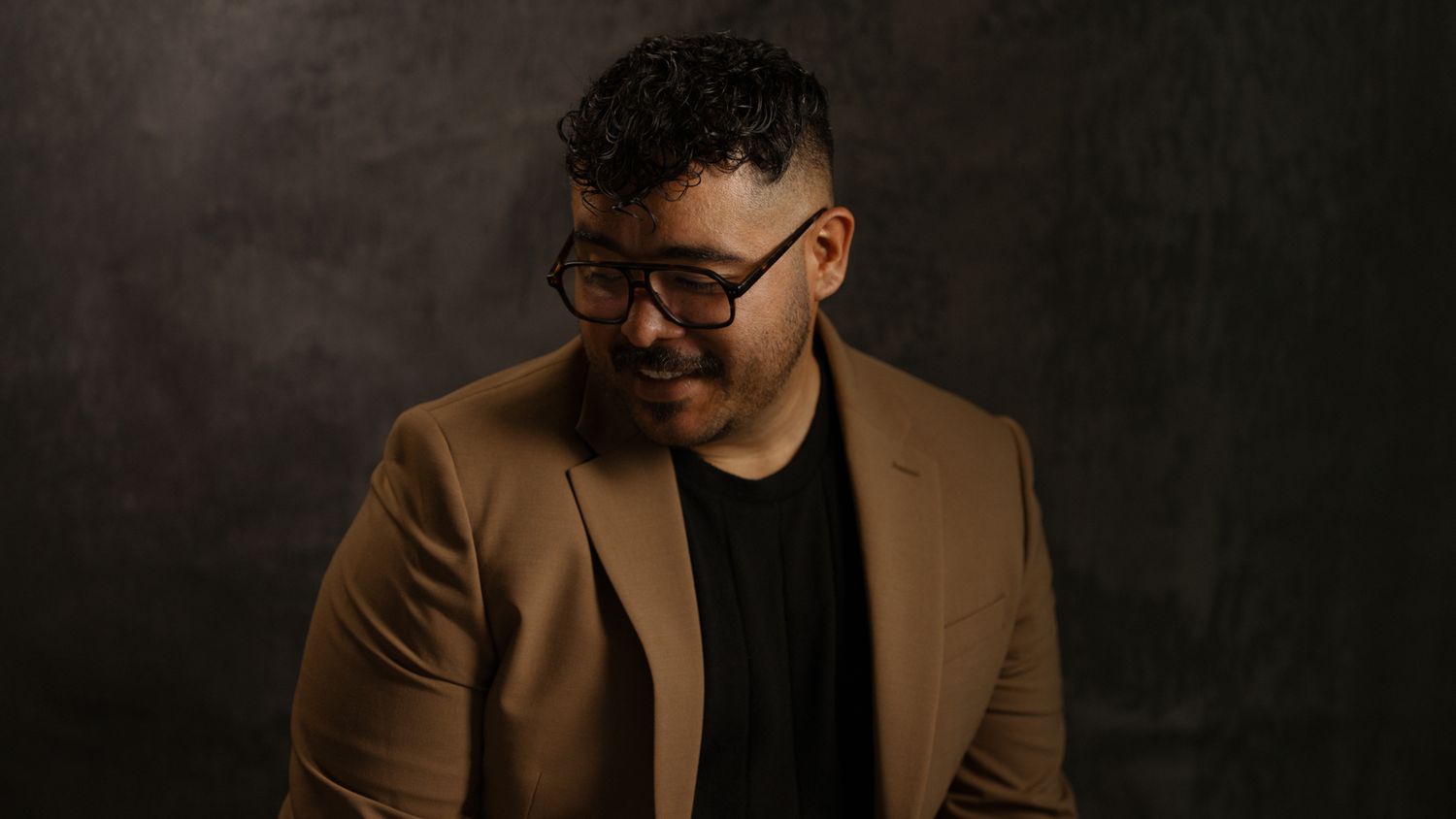 Man in tan blazer and black shirt smiling downward against dark textured background.