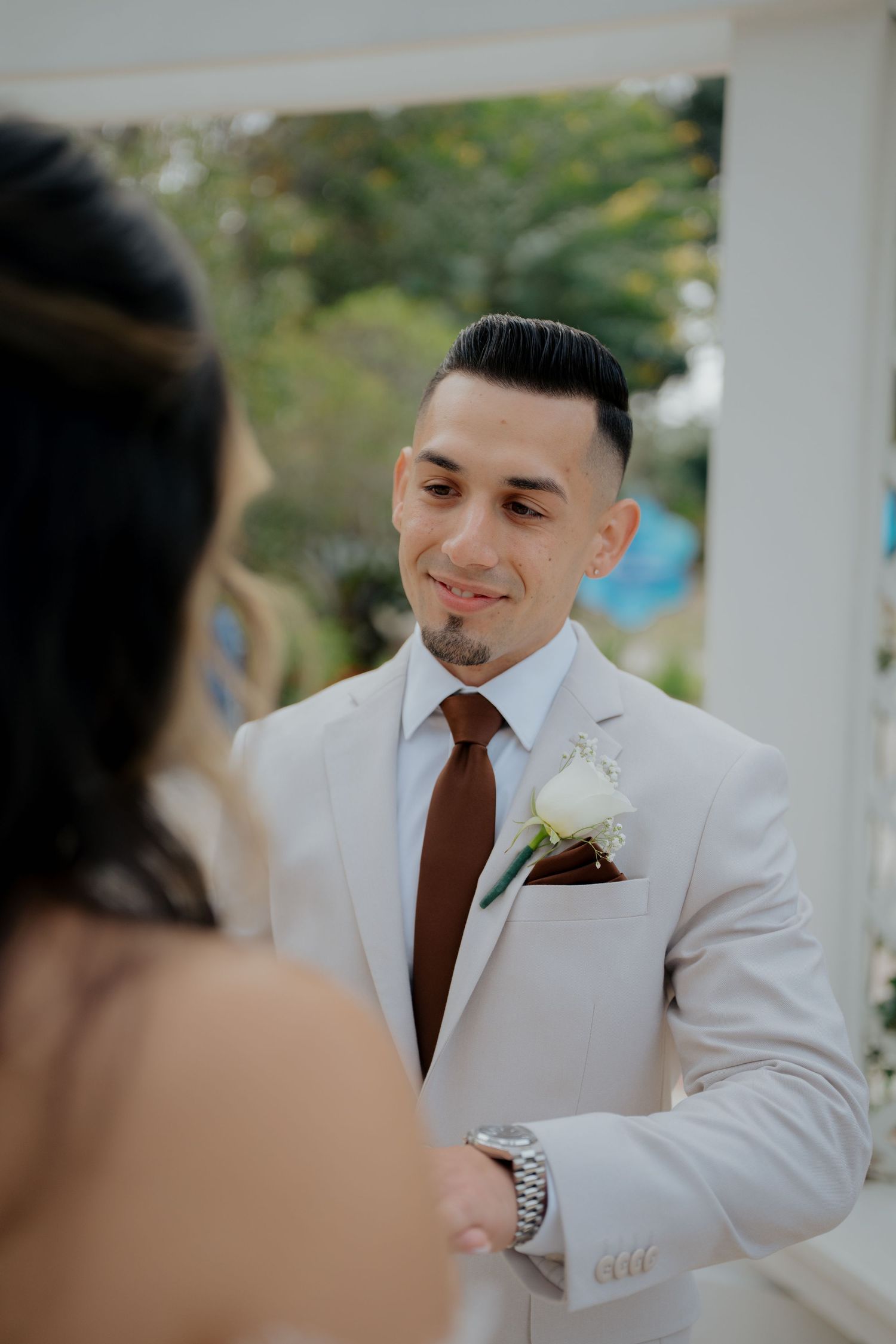 A groom in a white suit and brown tie smiles during an outdoor wedding ceremony.