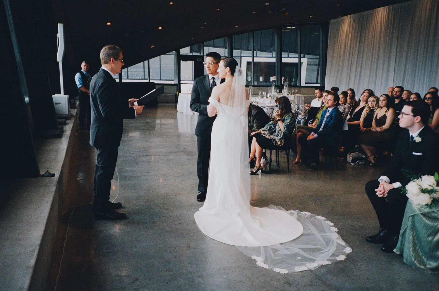 A couple stands at the altar during their modern wedding ceremony in an industrial-style venue with floor-to-ceiling windows.