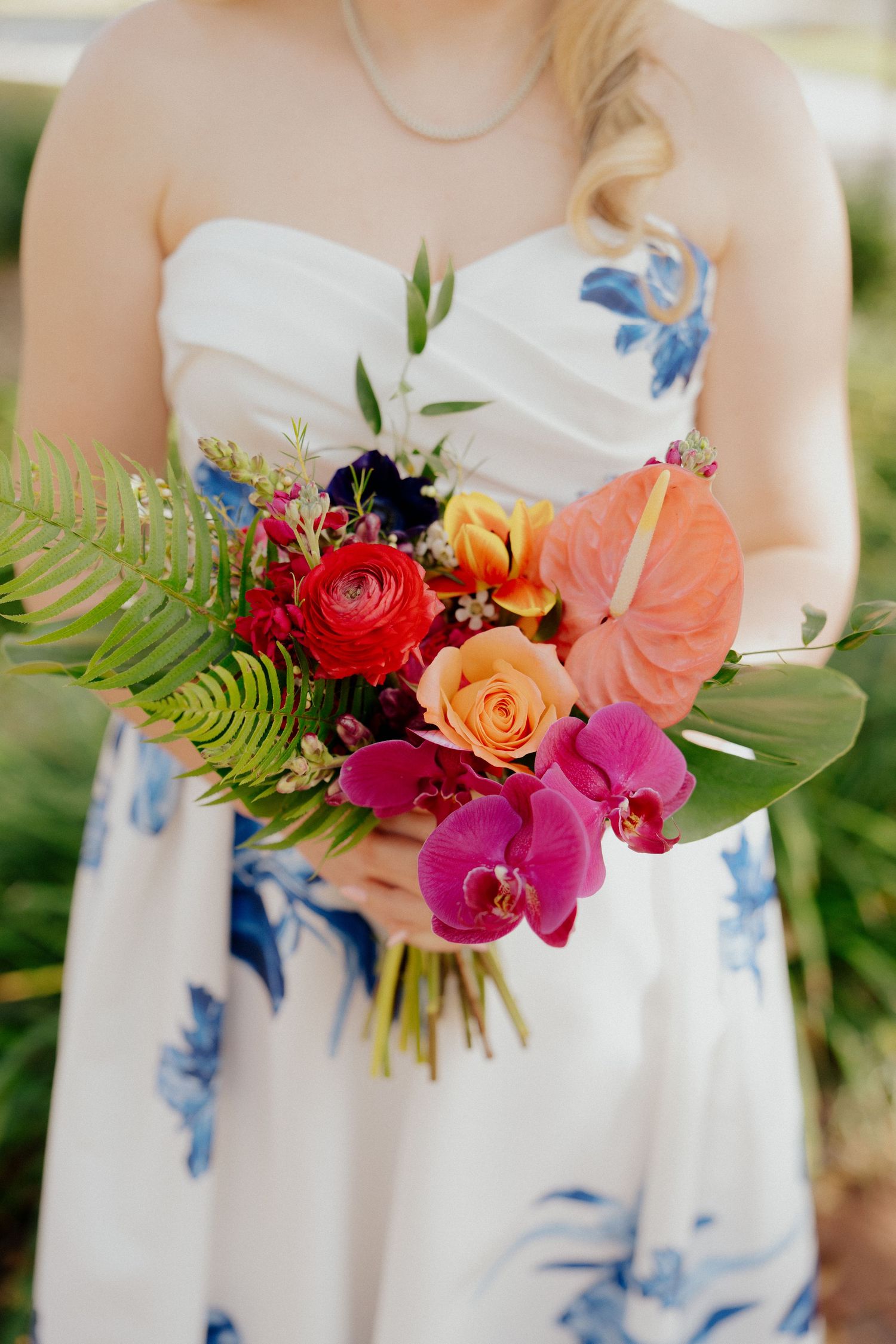 Bride holding a vibrant tropical wedding bouquet with pink orchids, orange roses, red ranunculus, and lush green ferns.