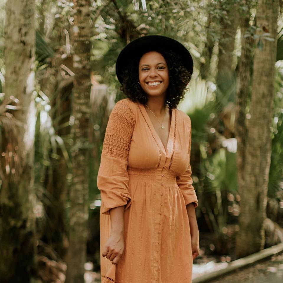 Smiling woman in orange dress and black hat standing among tropical green trees outdoors.