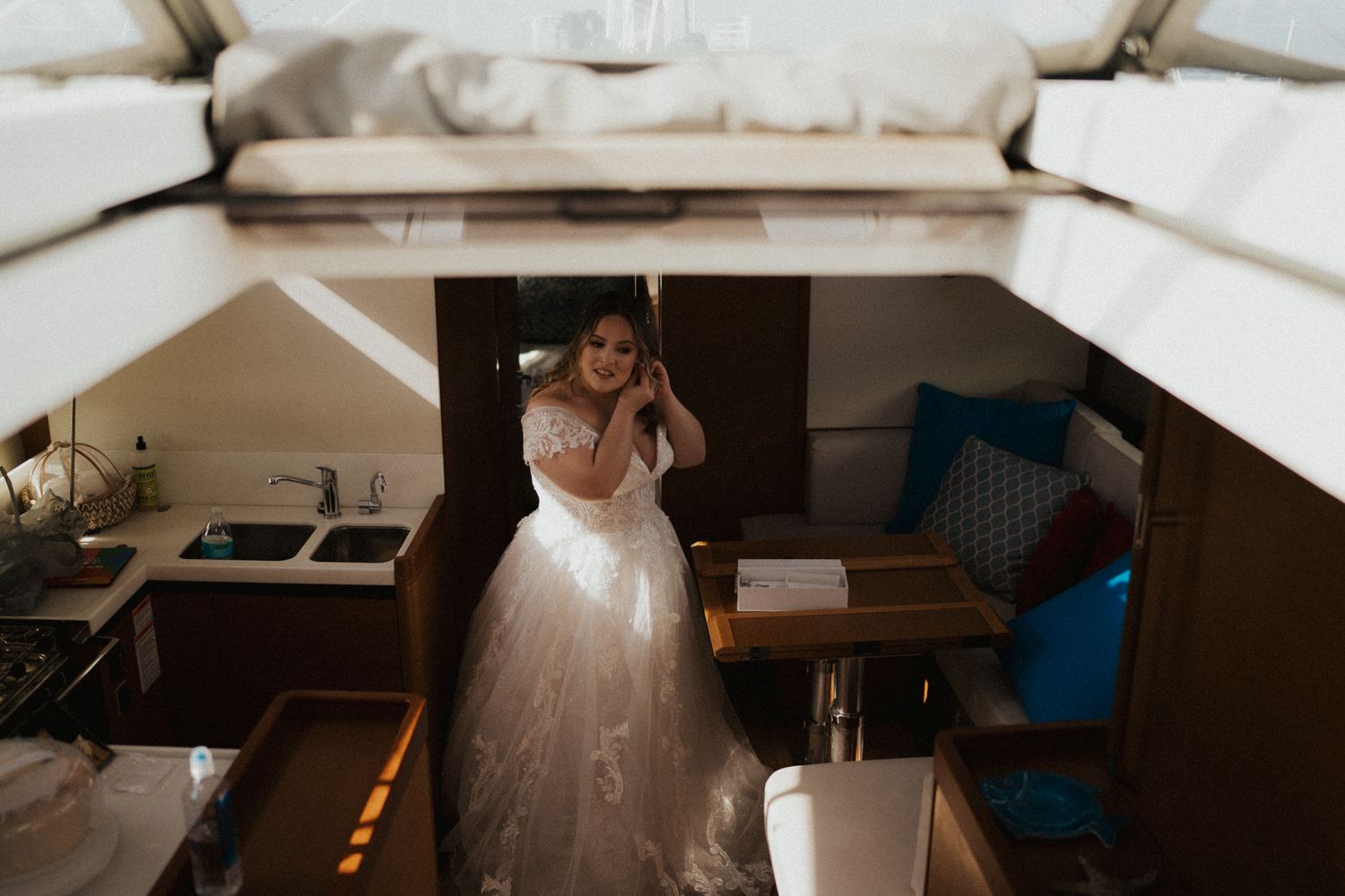 Bride in elegant white ball gown standing inside cozy sailboat cabin, viewed from above.
