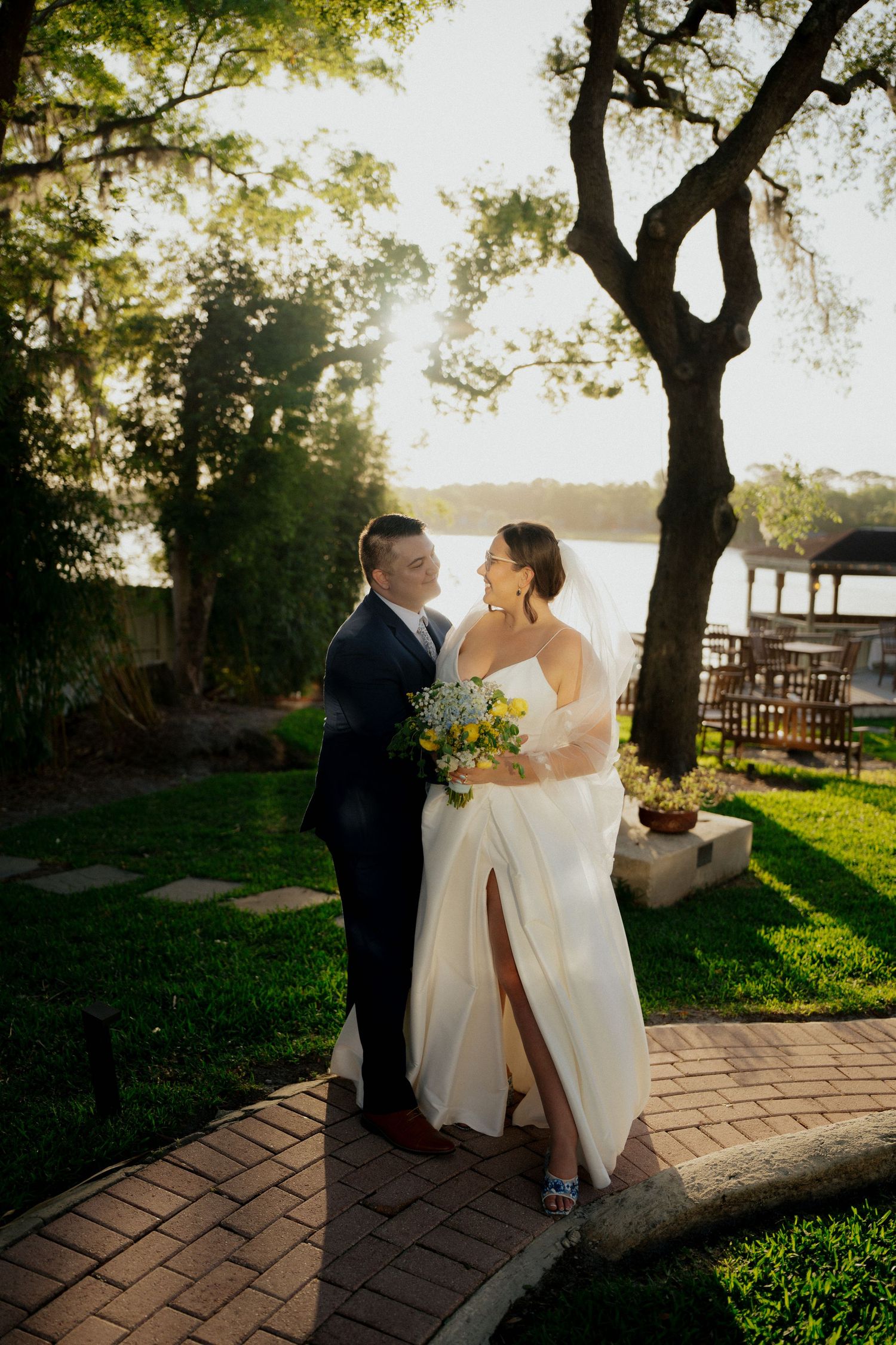Couple shares romantic moment at sunset on brick pathway near waterfront with tall tree and garden setting.