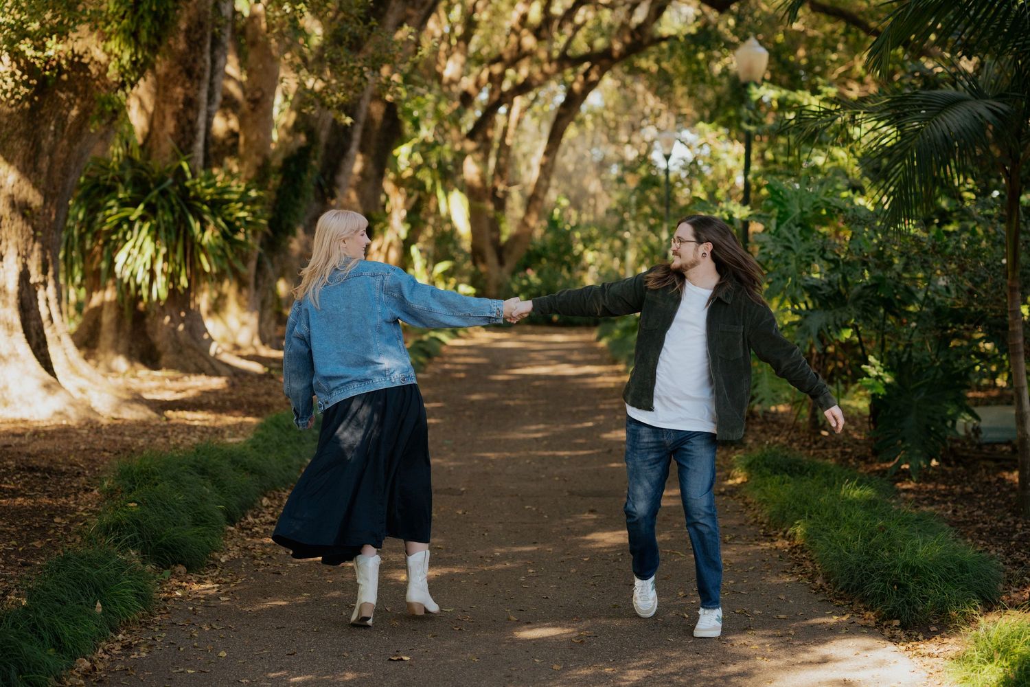 Two people share a playful moment on a tree-lined path in a park on a sunny day.