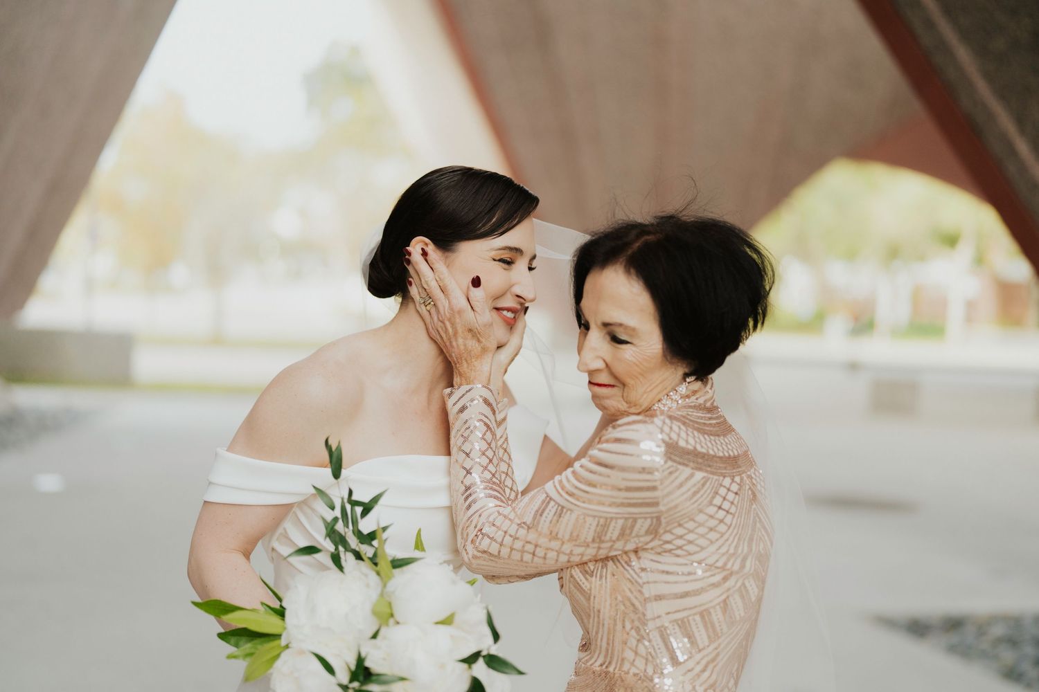 A tender moment between a bride and someone in a beaded blush dress holding white floral bouquets.