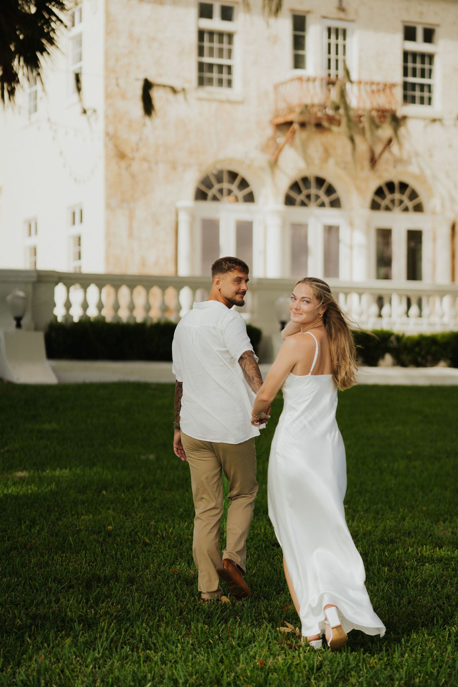 A couple in coordinated white and tan outfits poses in front of a Mediterranean-style mansion.