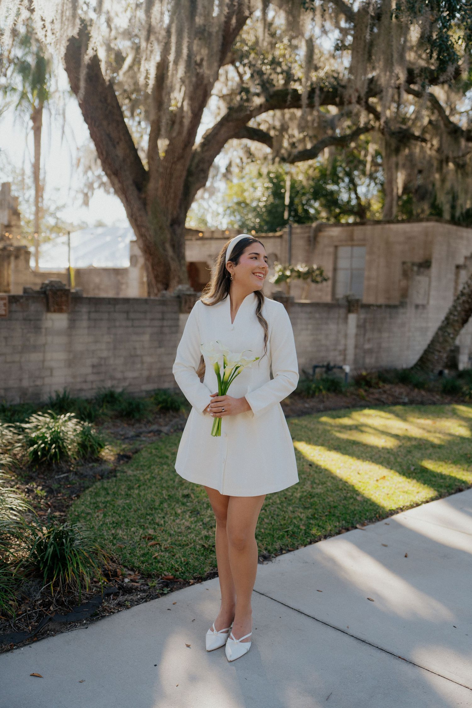 A person in a white mini dress and sneakers poses outdoors under a Spanish moss draped tree.