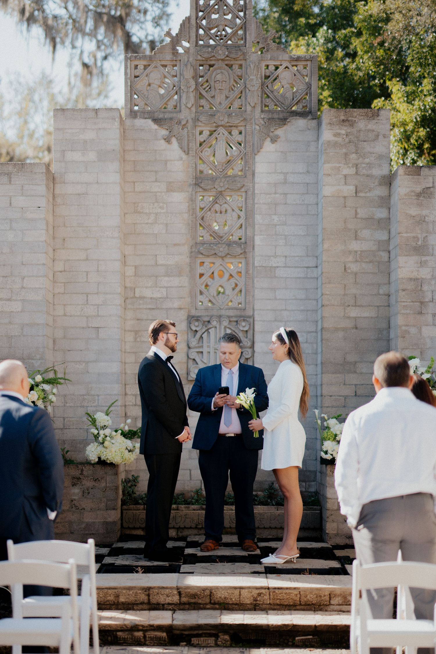 Small intimate outdoor wedding ceremony with white chairs and decorative stone wall backdrop.