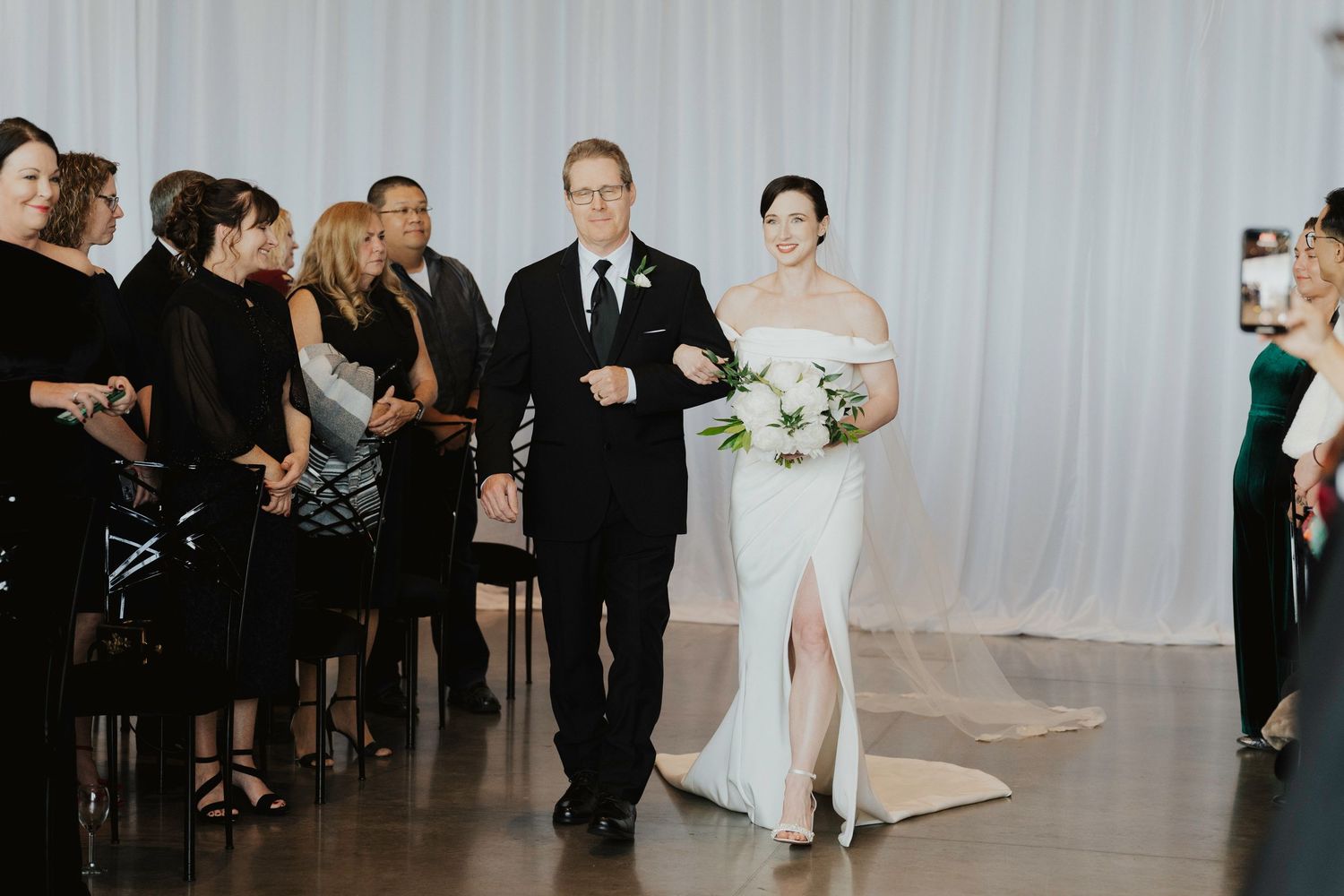 A wedding ceremony processional with guests watching as a couple walks down the aisle against a white backdrop.