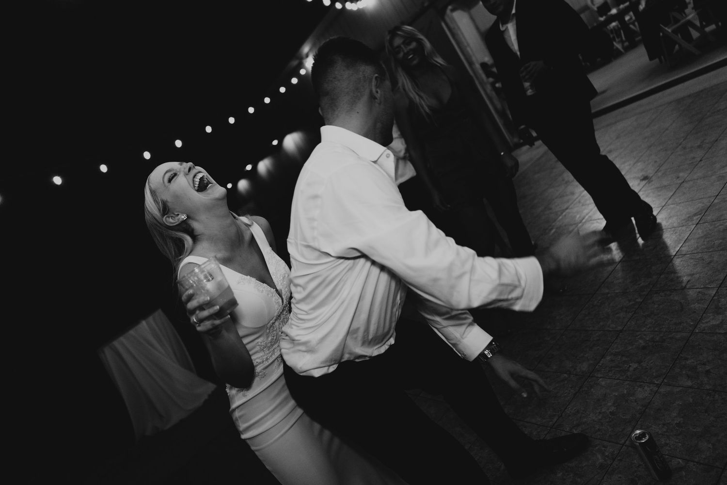 Black and white photo of a couple dancing joyfully at a wedding reception under string lights.
