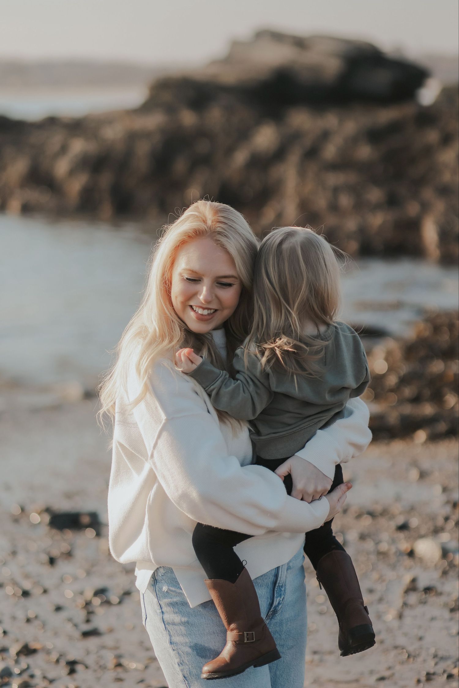 A smiling blonde woman holds a young child on a rocky beach at golden hour.