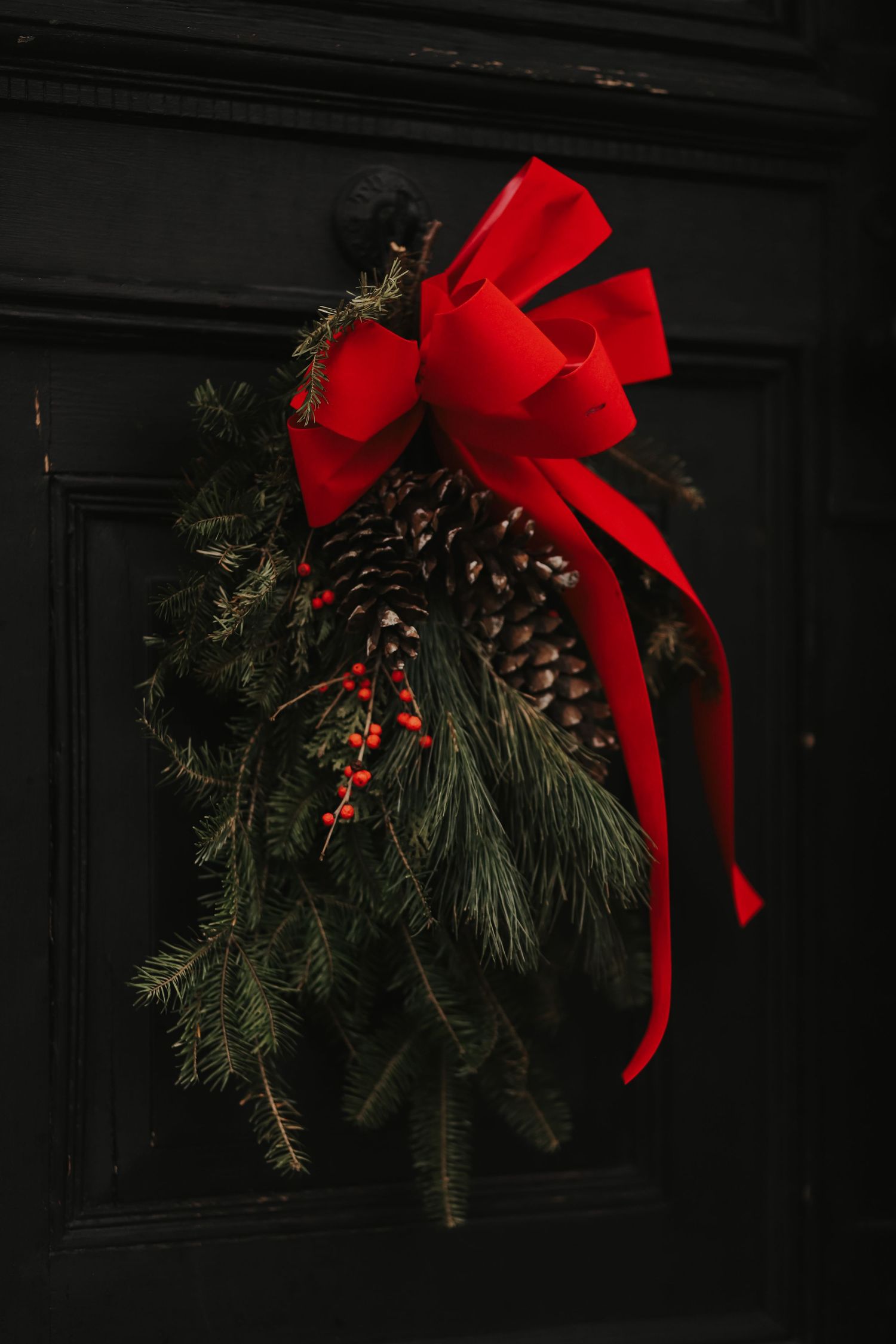 Festive Christmas door swag with pine branches, pinecones, red berries, and a red bow on black door.