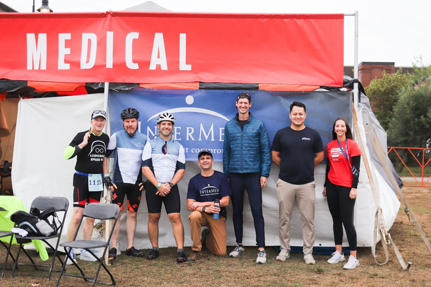 Group of seven people posing under a red Medical tent at an outdoor cycling event.