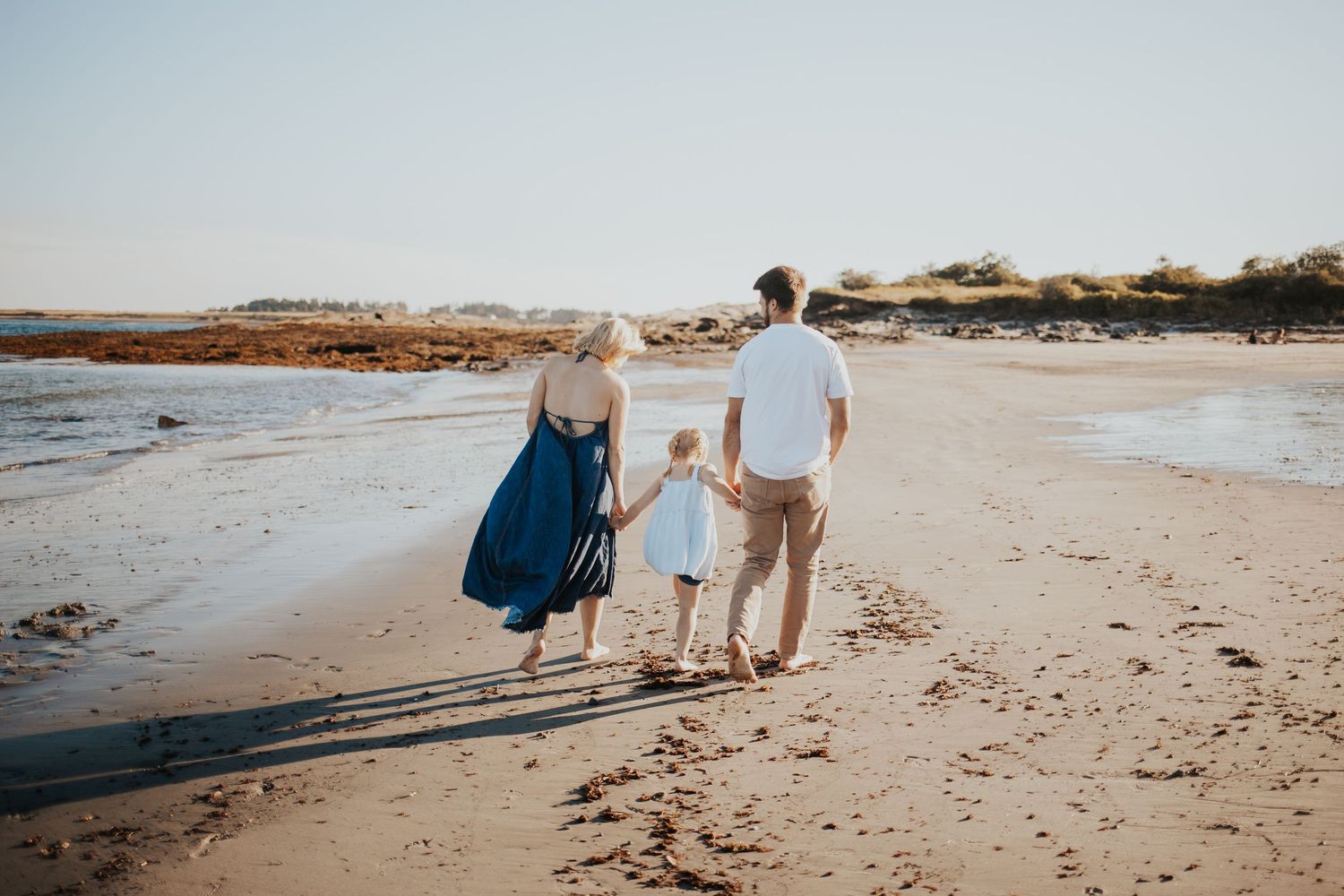 Family of three walking hand-in-hand along a sunny beach, mother in blue dress, father in white shirt, child in white dress.
