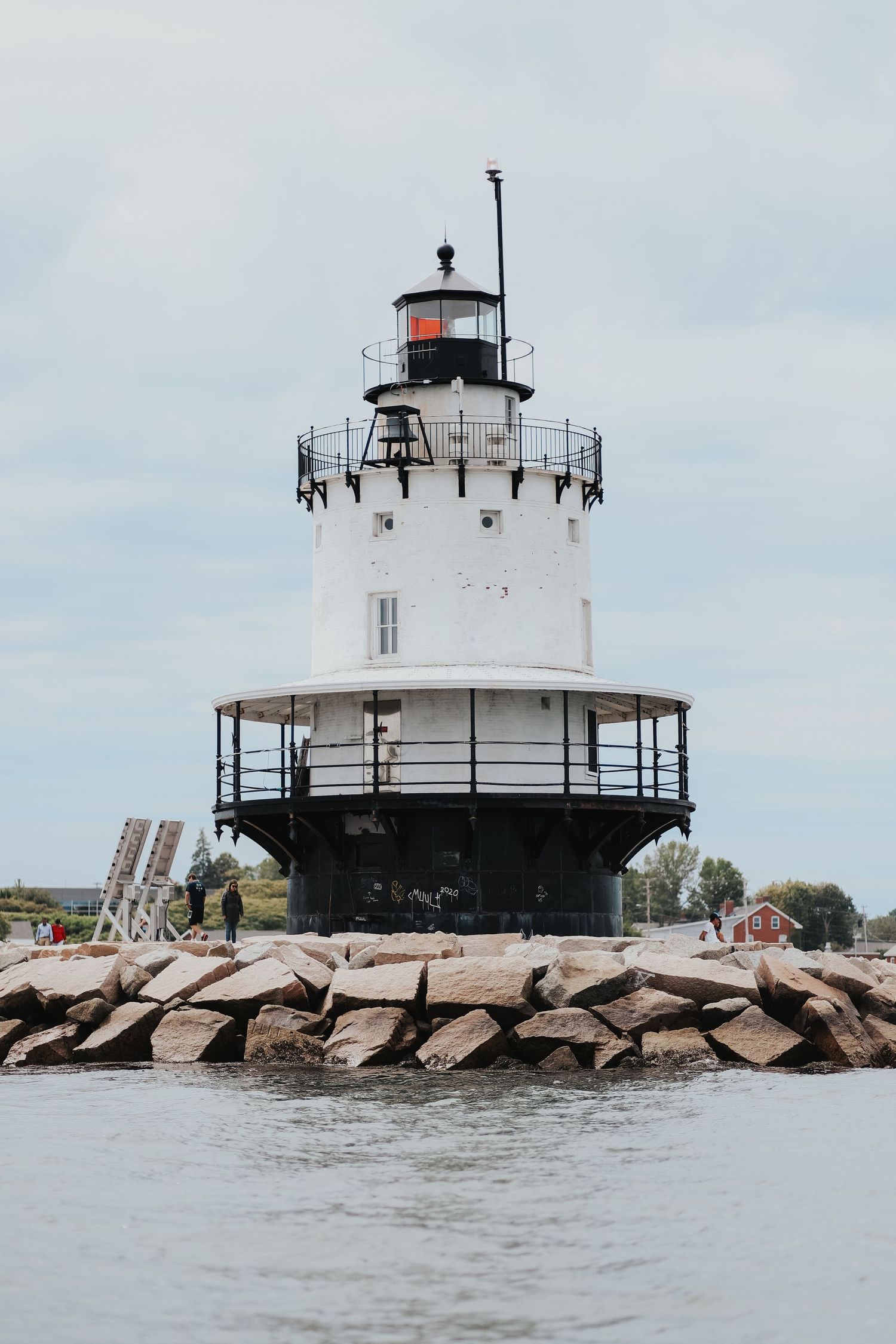 White cylindrical lighthouse on rocky breakwater with metal walkway, overcast sky, and calm harbor waters in background.