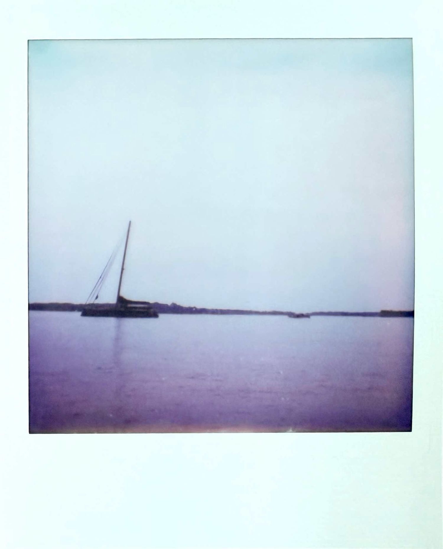 Lone sailboat on calm water with muted purple sky and distant shoreline, vintage Polaroid photo.