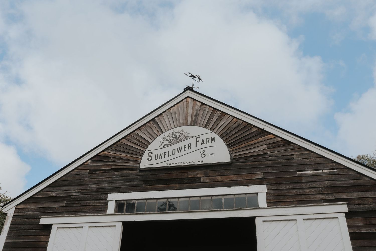 Rustic wooden barn with Sunflower Farm sign on gable against blue cloudy sky.
