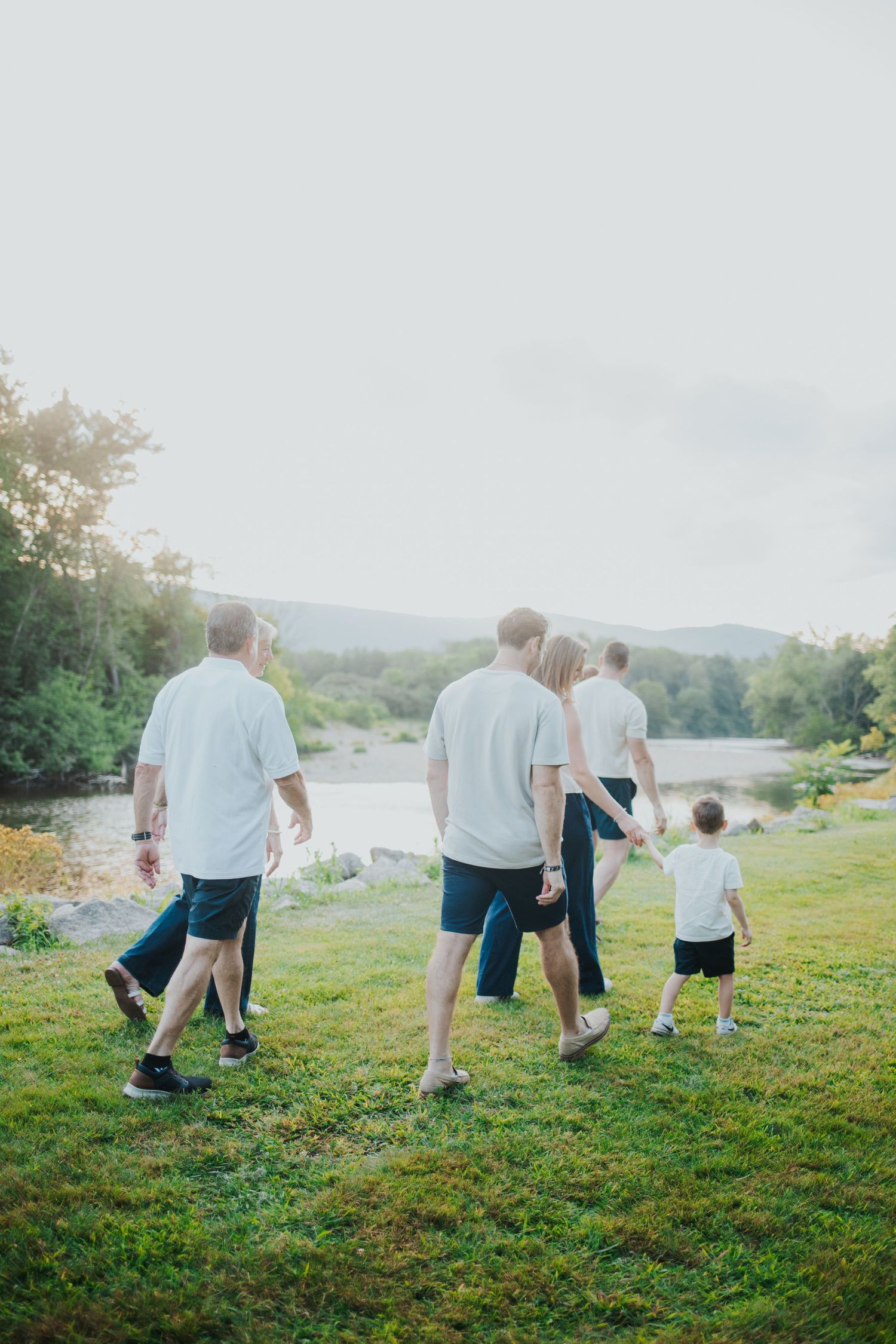 Family in white shirts walking on green grass near a river during golden hour sunset.