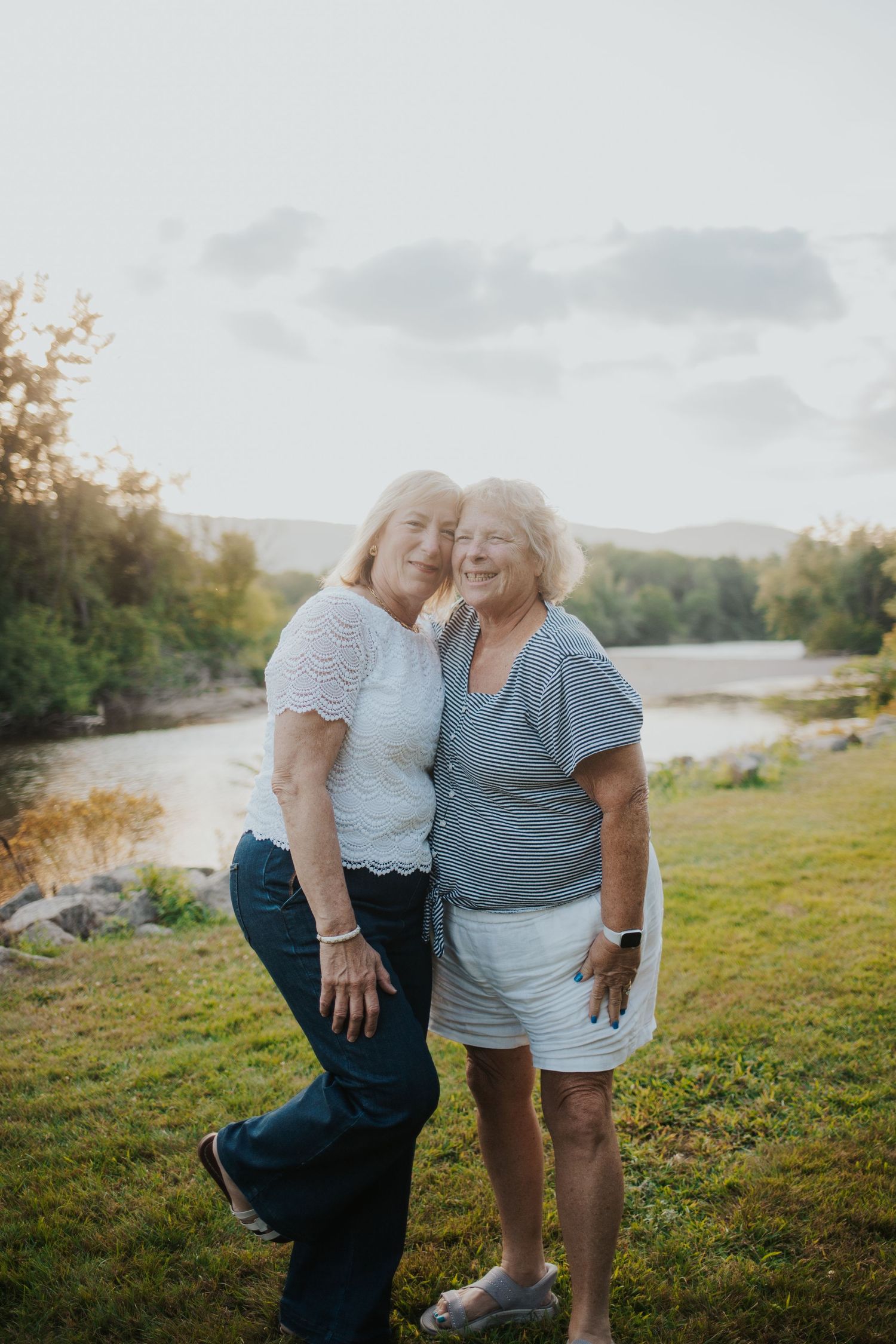 Two older women smiling and embracing outdoors near a river with trees and mountains behind them.