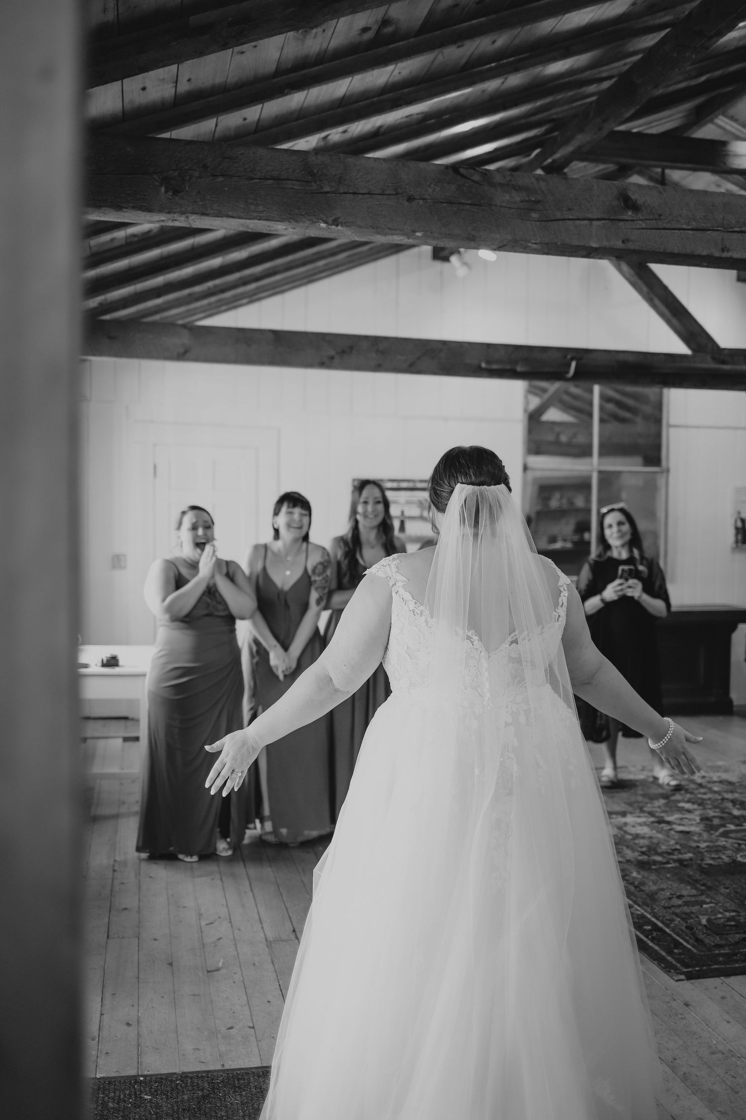 Black and white photo of bride in wedding gown and veil facing emotional bridesmaids in a rustic barn venue.
