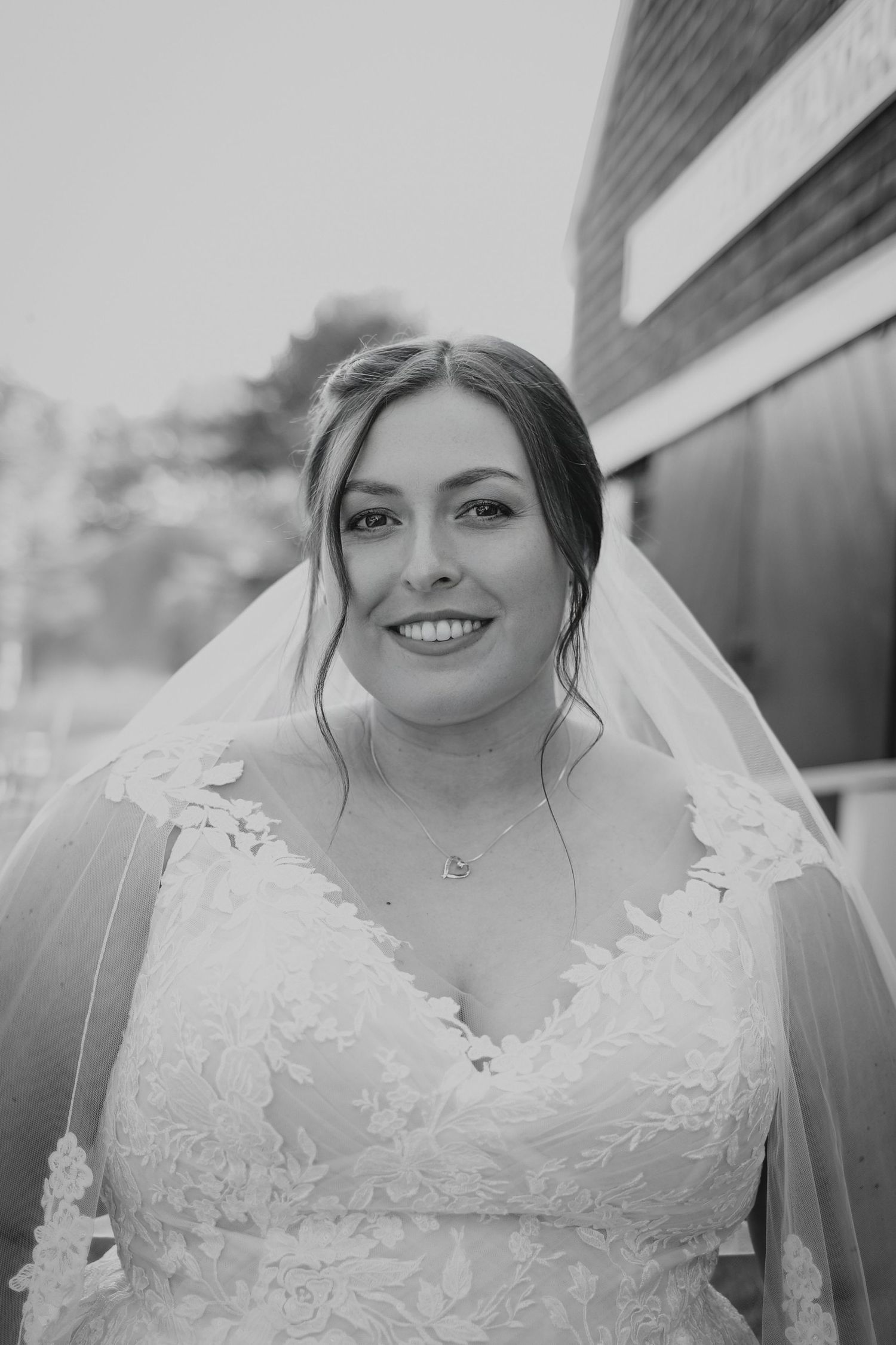 Black and white bridal portrait of smiling bride in lace wedding gown and veil outdoors.