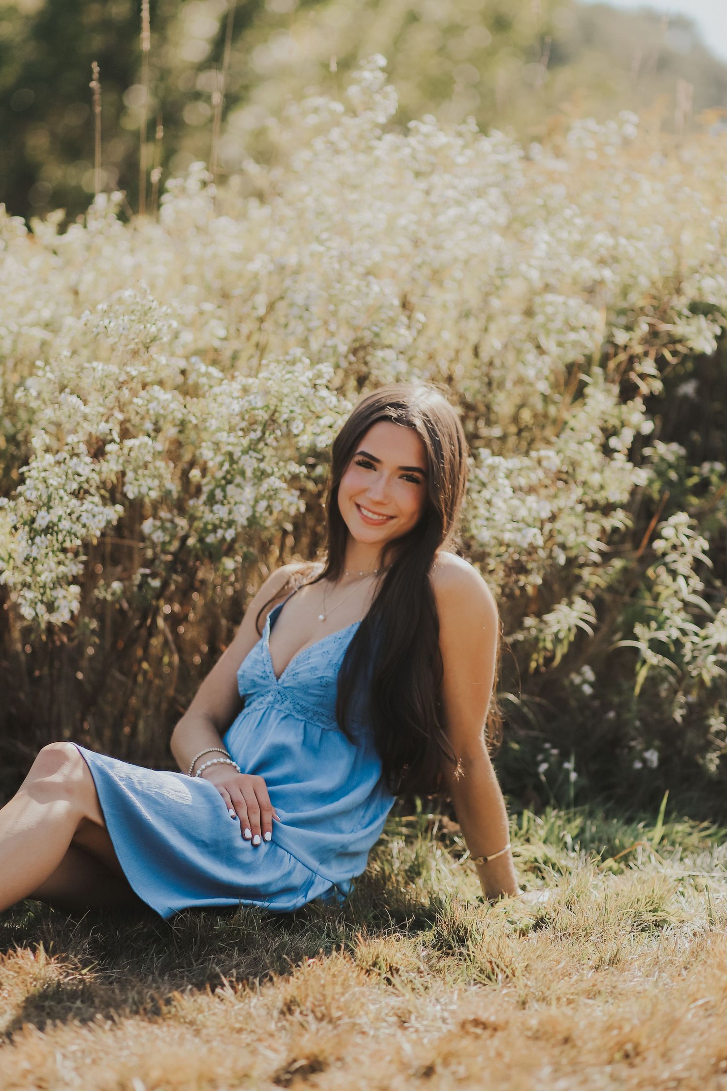 Young brunette woman in blue sundress sitting in golden field of wildflowers smiling at camera.