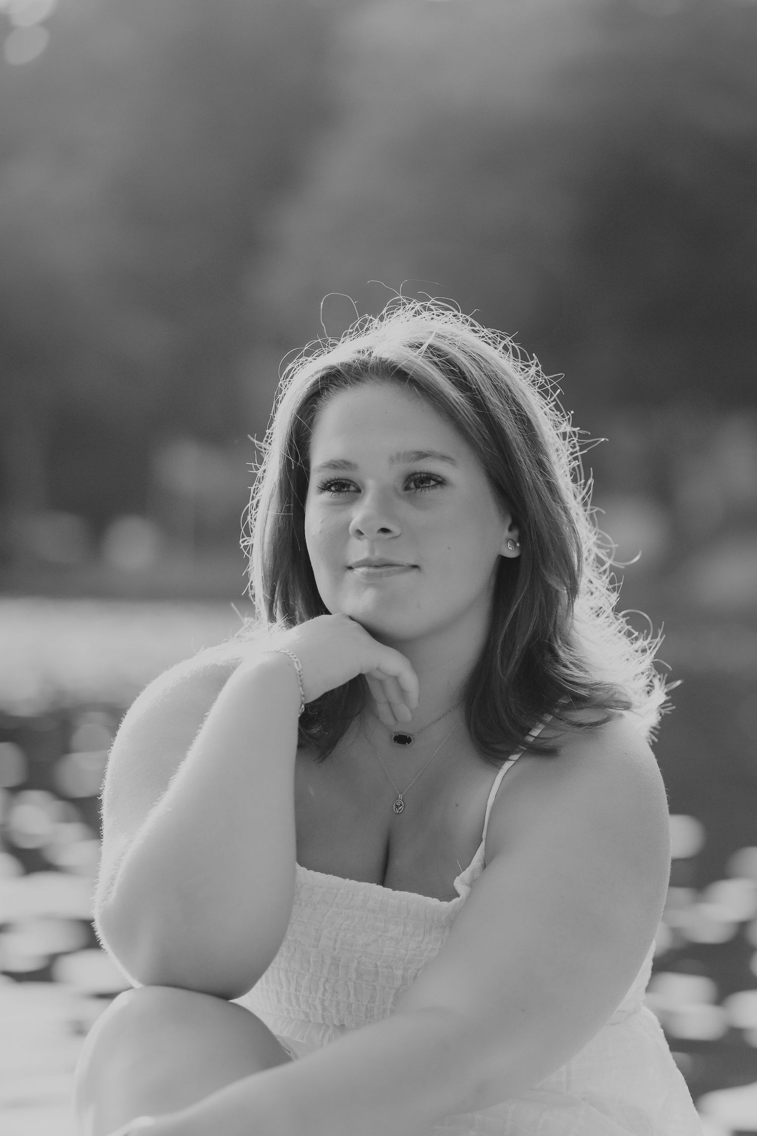 Black and white portrait of a young woman resting her chin on her hand near a sunlit pond.