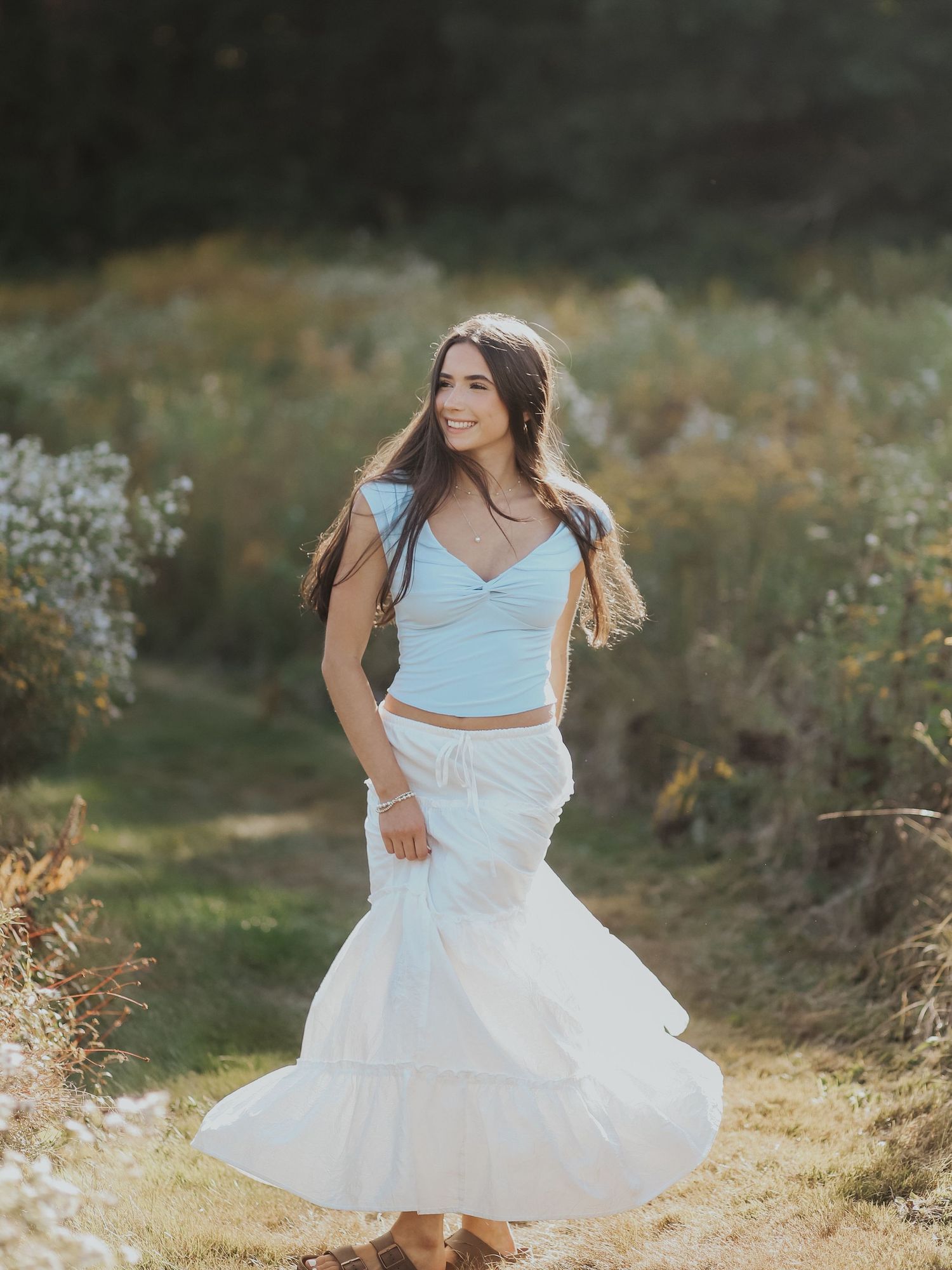 Young woman in light blue crop top and white tiered maxi skirt twirling outdoors in golden sunlight.