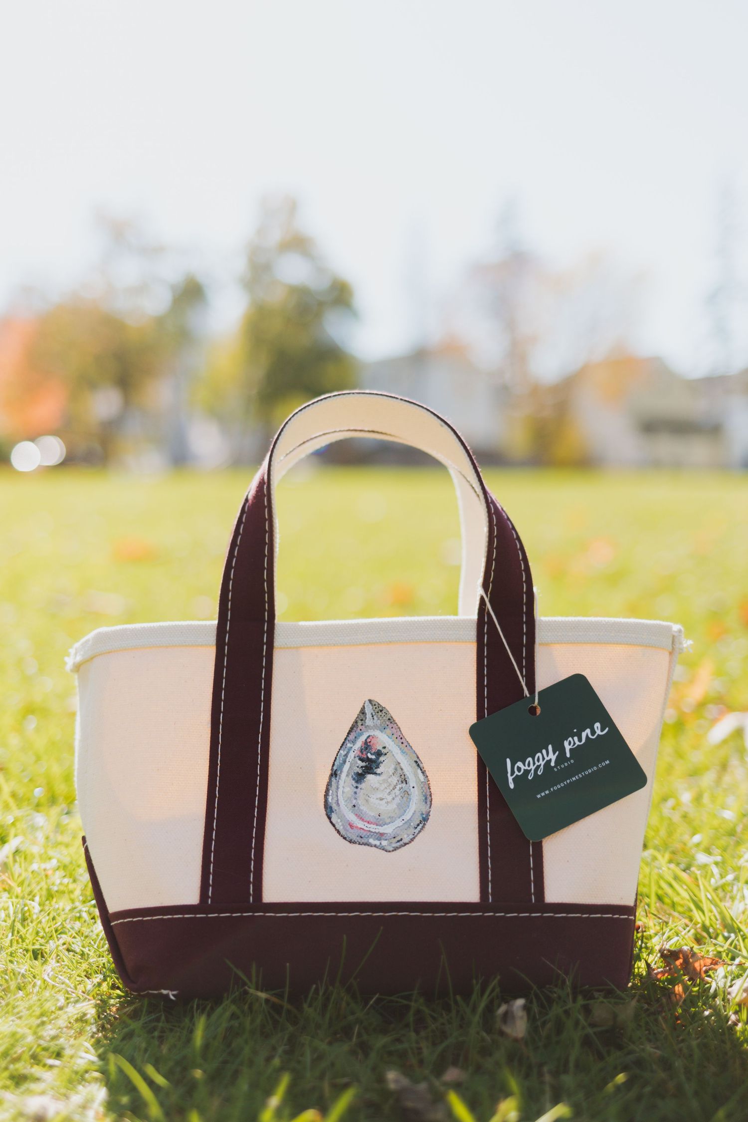 Cream canvas tote bag with maroon handles and trim, featuring a painted oyster design and a Foggy Pine tag, resting on sunlit green grass.