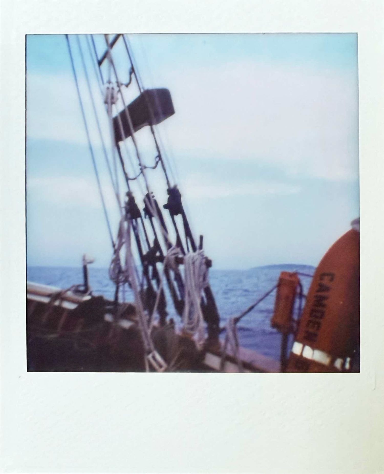 Polaroid photo of tall ship rigging and mast against a blue sky with an island in the distance.