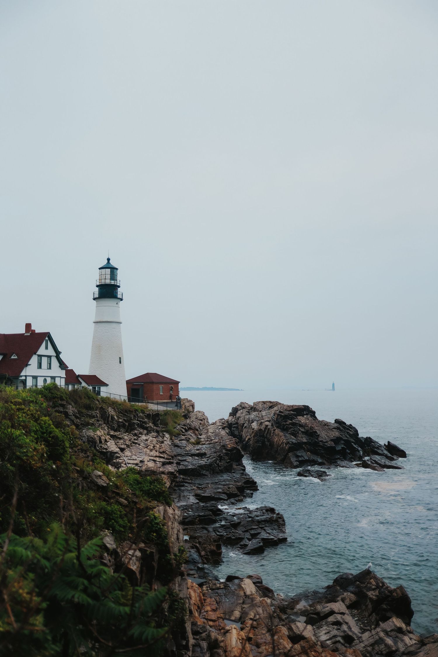 White lighthouse with a black lantern room and red-roofed buildings on a rocky coastal cliff overlooking the calm sea under a hazy gray sky.