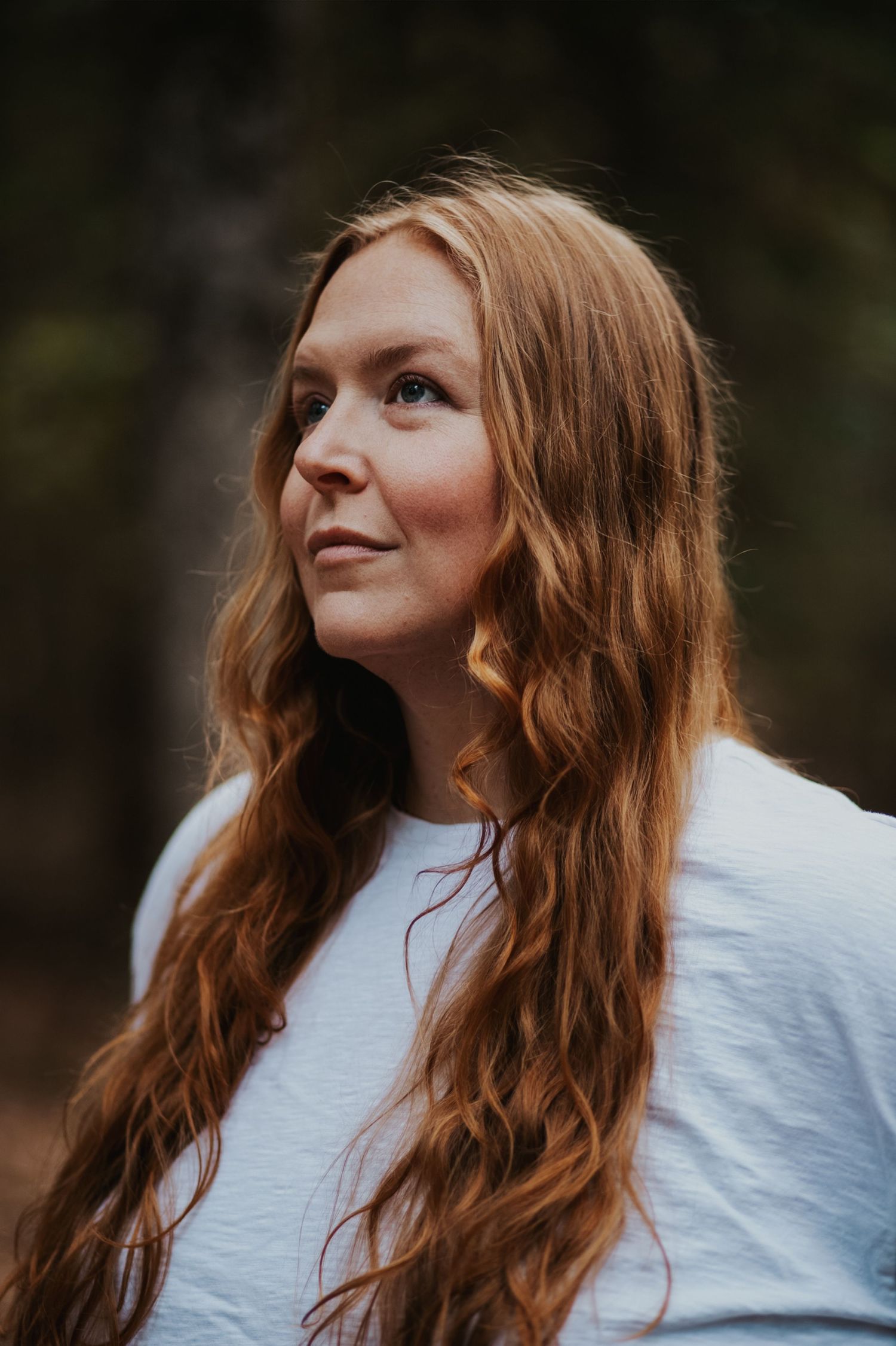 A young woman with long wavy red hair gazes upward outdoors, wearing a white shirt with a blurred forest background.