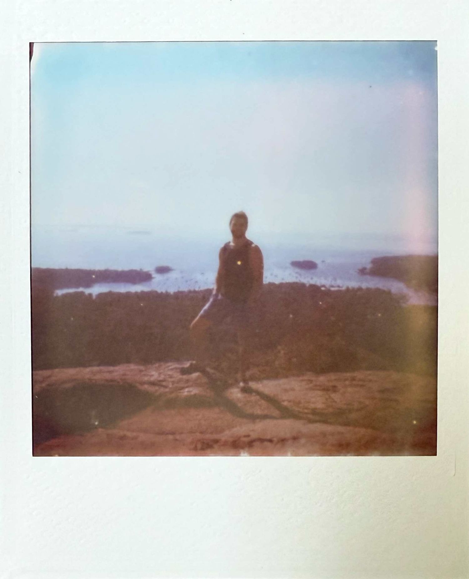 Person standing on rocky coastal overlook with ocean islands visible in the distance, vintage Polaroid photo.