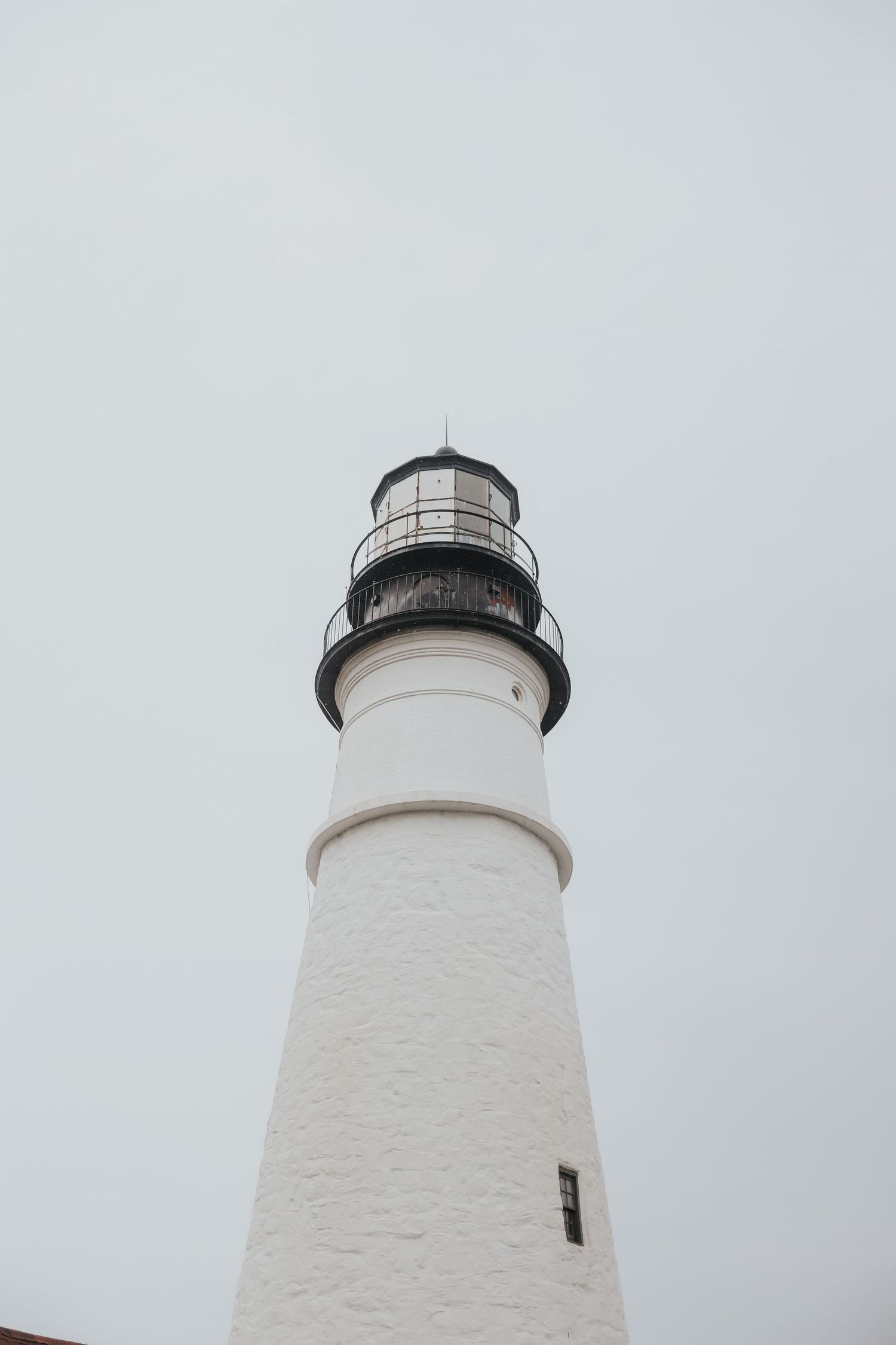 White lighthouse tower photographed from below against overcast sky, showing textured exterior and black lantern room at top.