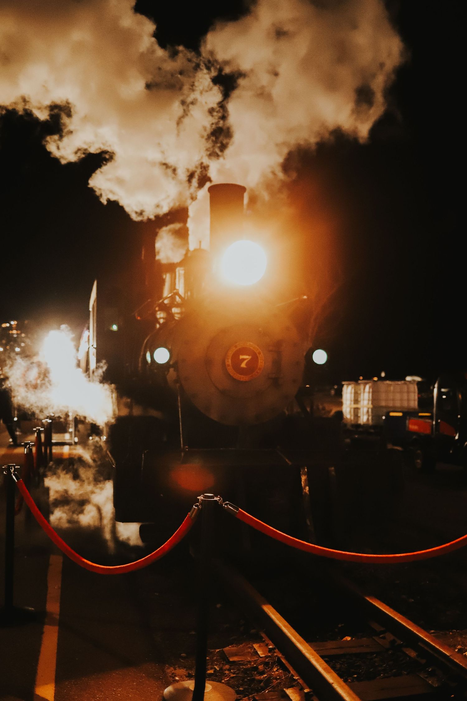 Antique steam engine number 7 glowing at night with smoke and red rope barriers on tracks.
